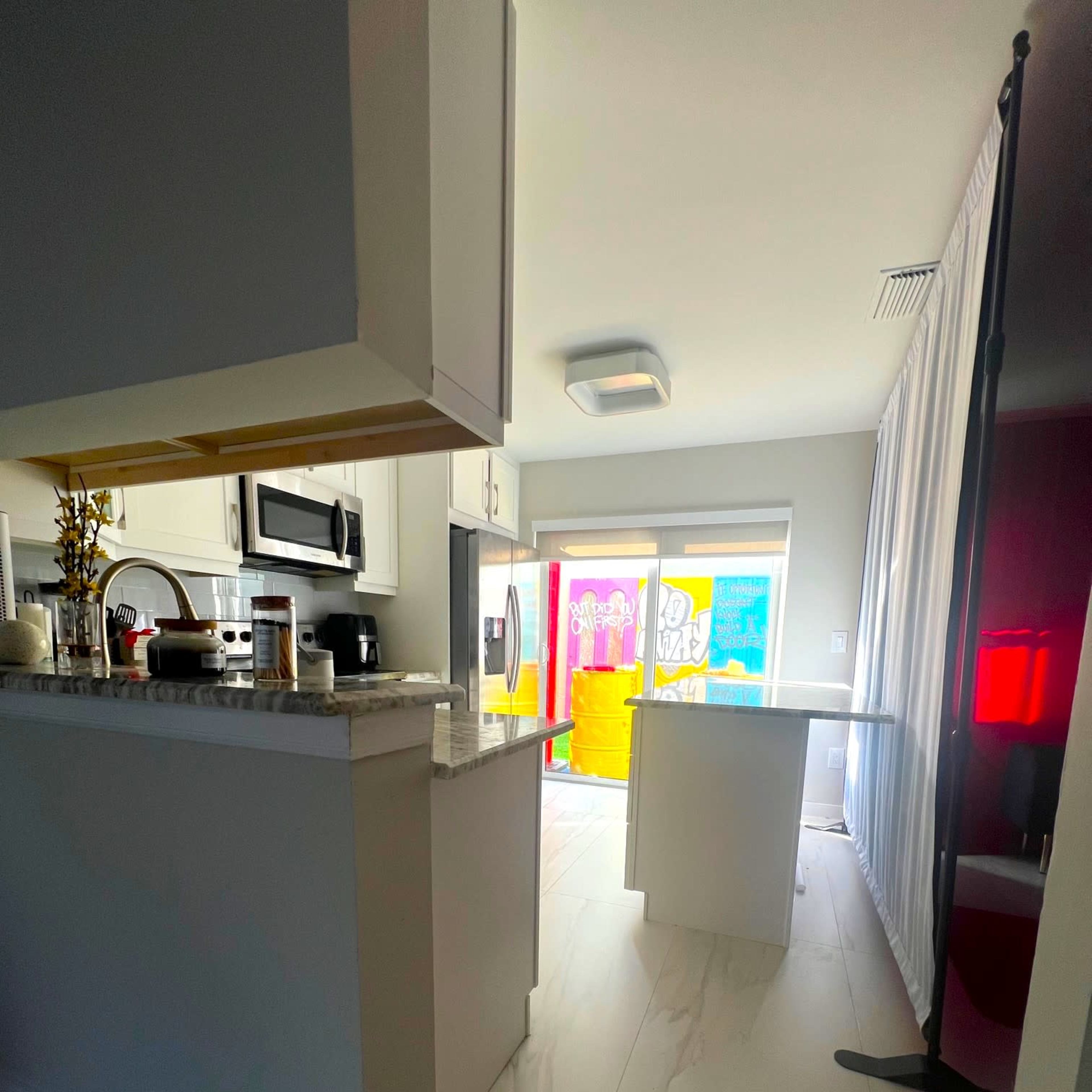 A bright kitchen features white cabinetry and a granite countertop, with a view of colorful structures outside through a large window.