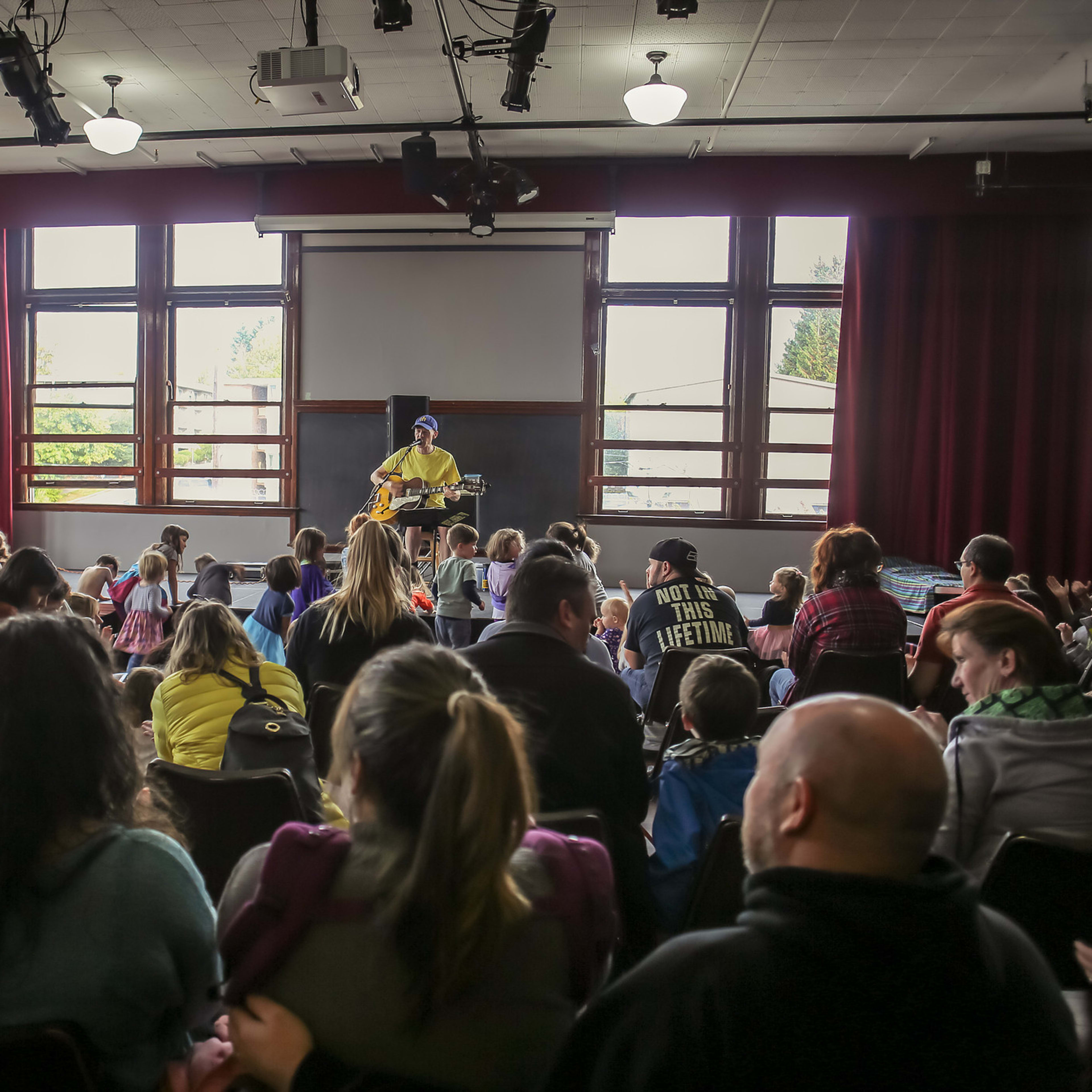 A performer plays guitar on a small stage in front of an audience seated in a room with large windows.