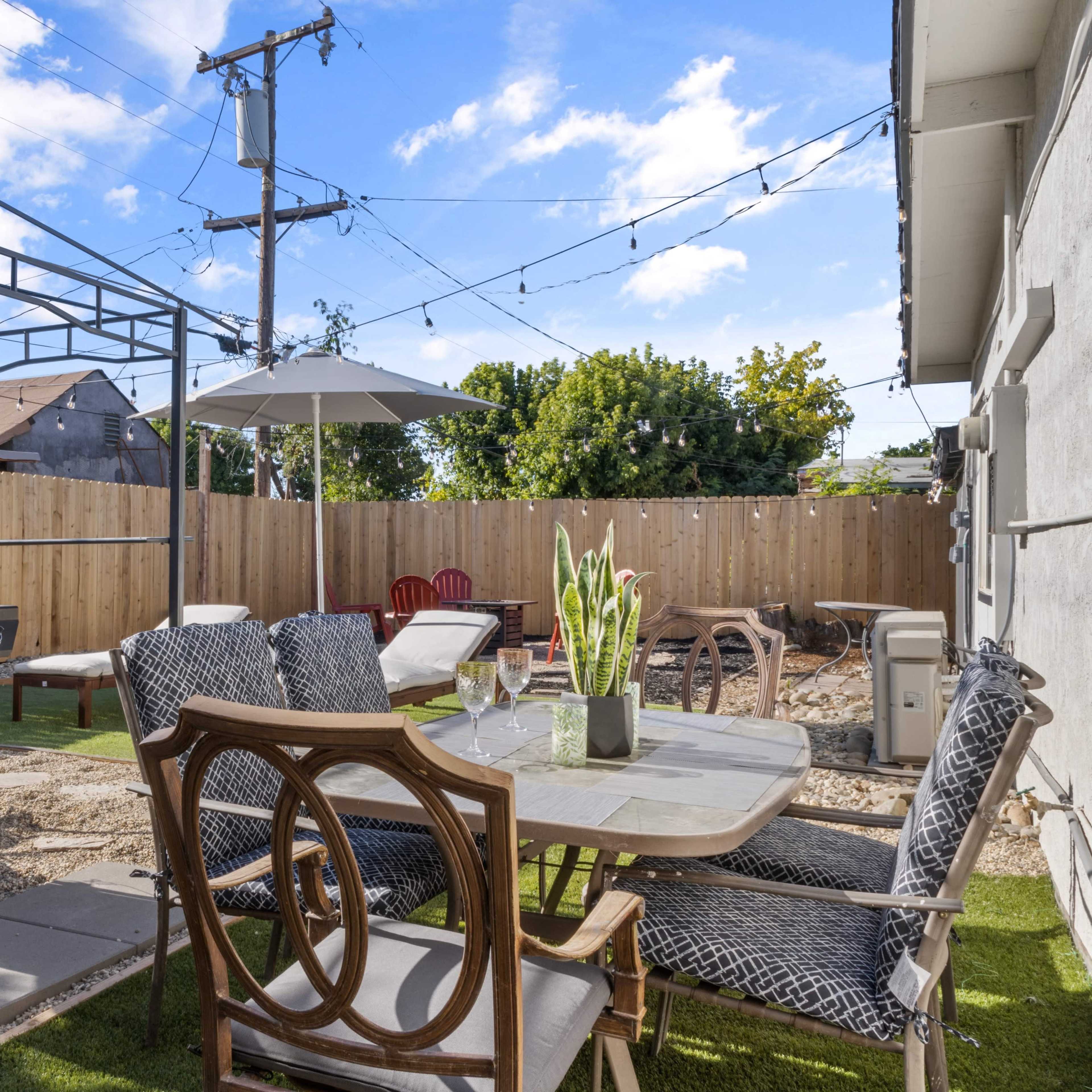 A backyard patio area with a grill, dining table, and chairs, surrounded by a wooden fence and string lights.