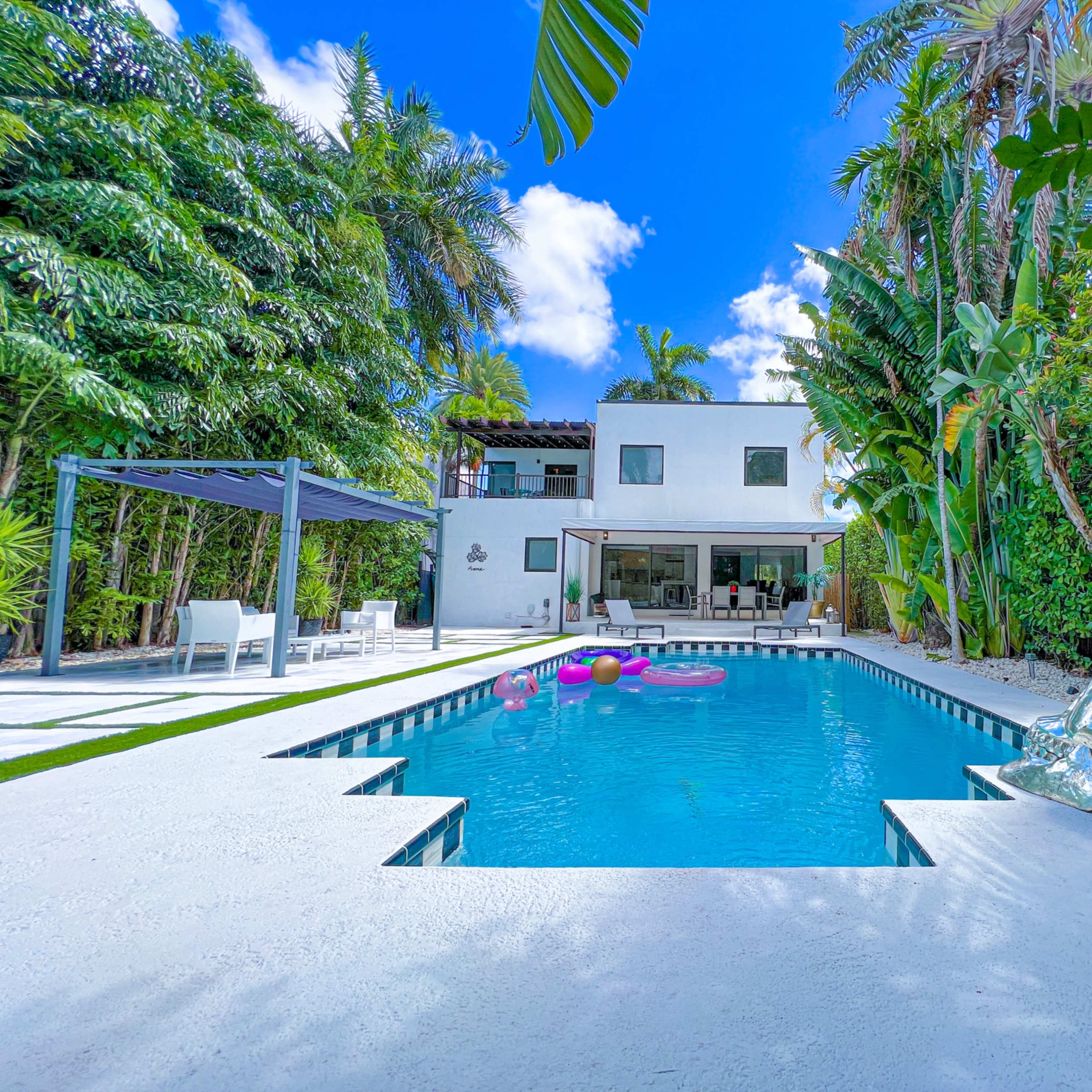 A modern house with large windows overlooks a swimming pool surrounded by tropical plants and a decorative silver skull sculpture.