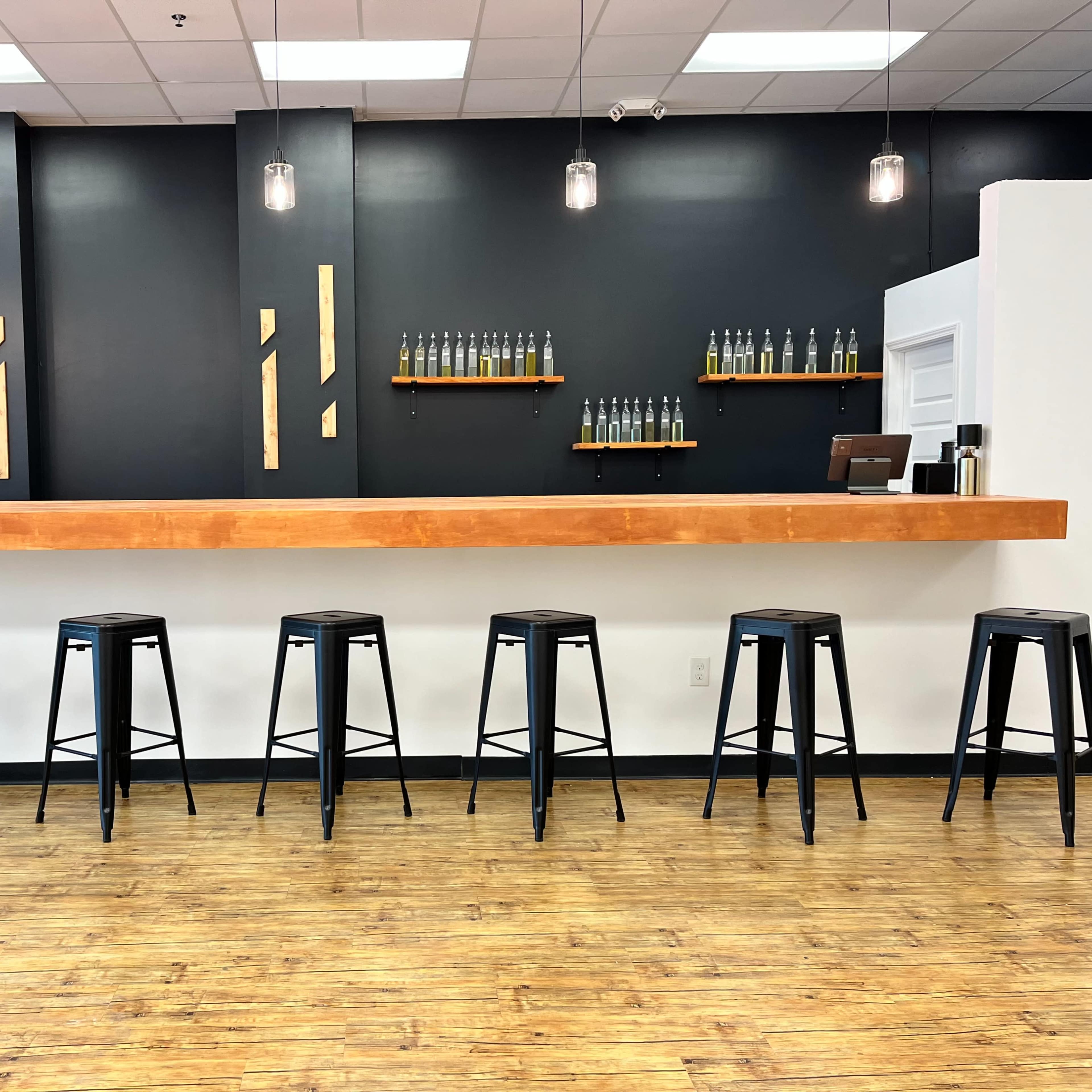 A minimalist bar area with a wooden countertop and several black stools lined up in front of a dark wall, featuring shelves with bottles on display.
