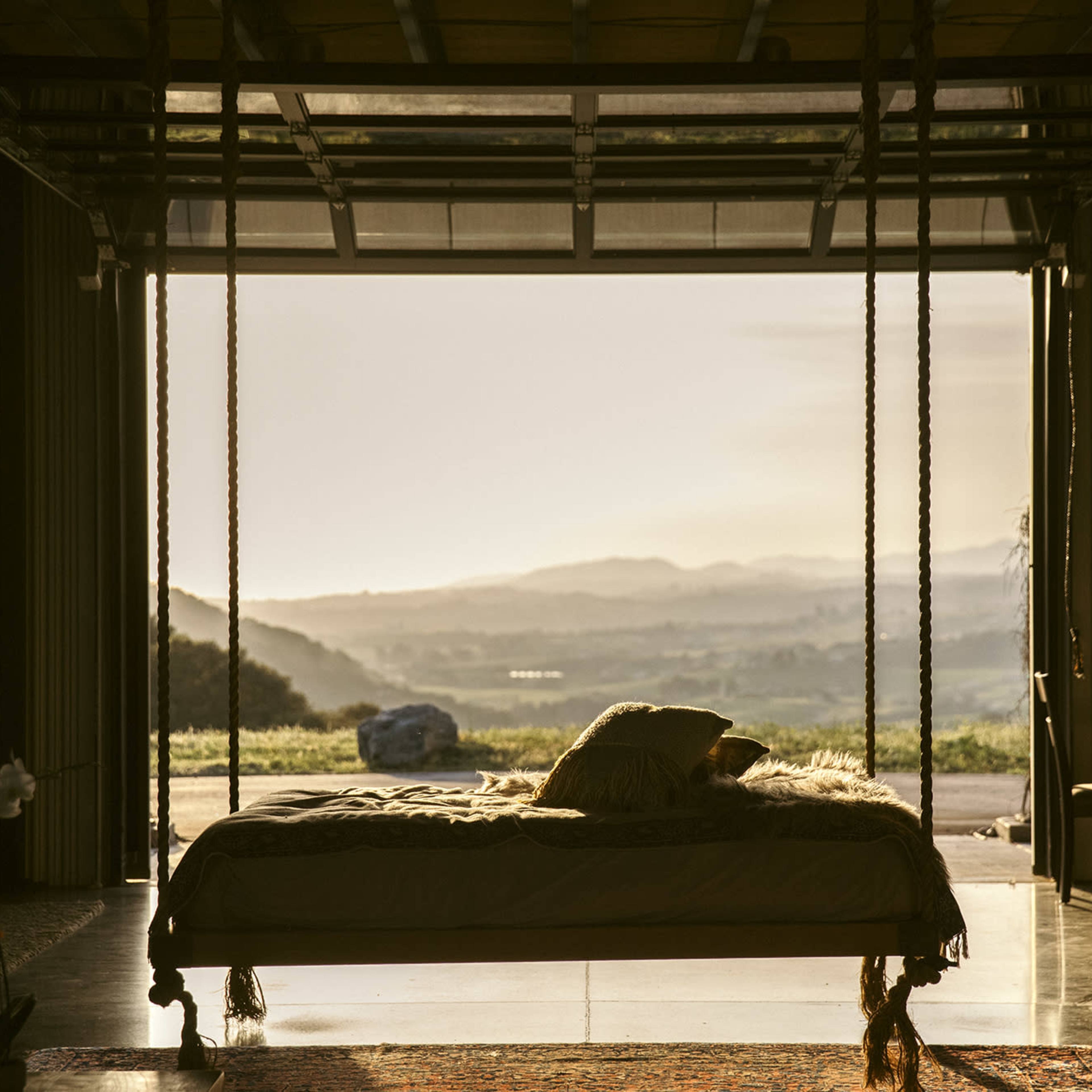 A bed is placed in an open space with glass doors that provide a view of a mountainous landscape at sunset.