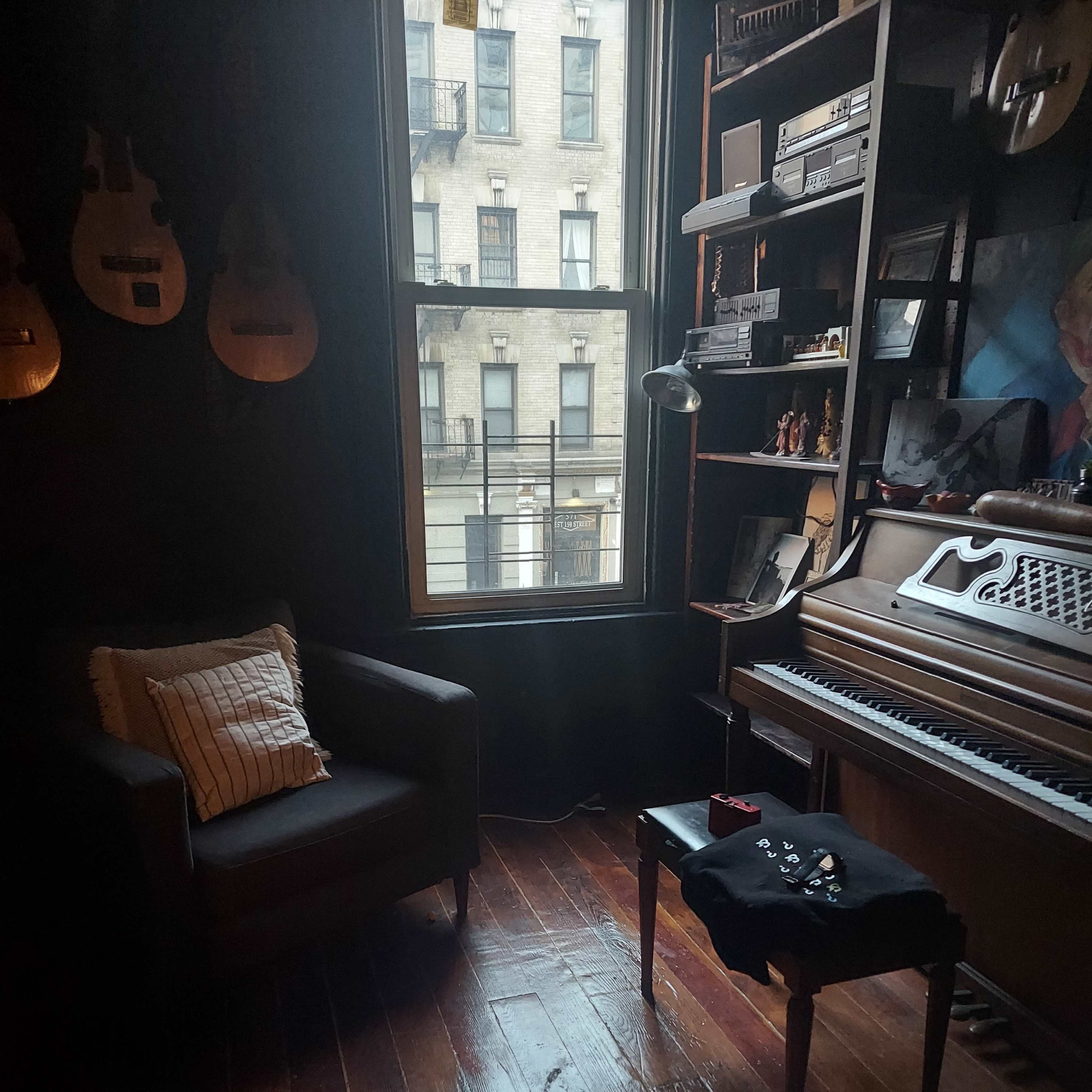 A cozy corner of a room features a chair next to a wooden piano and a small table with items on it, illuminated by natural light from a nearby window.