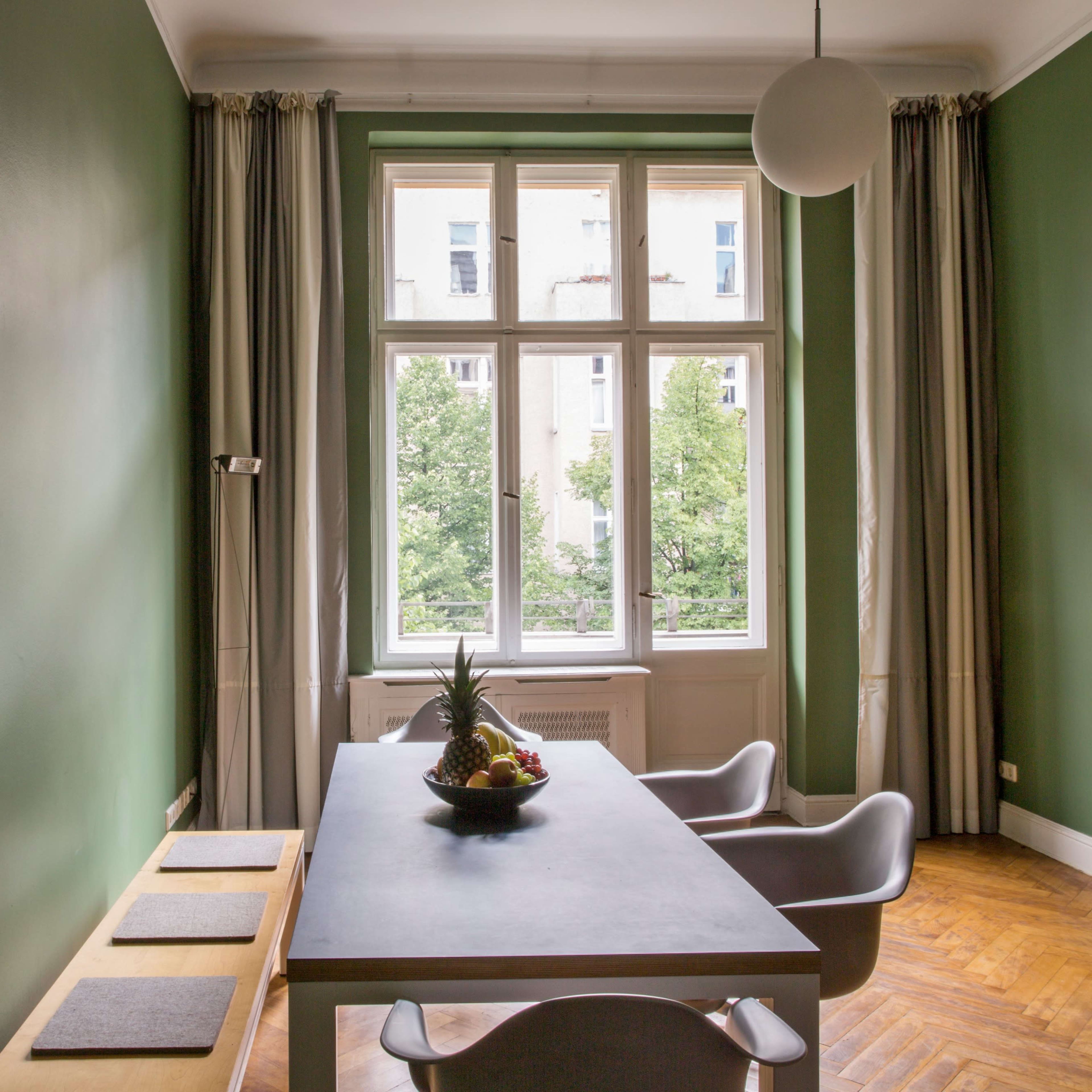 A dining area features a table with a fruit bowl, surrounded by modern chairs and large windows overlooking a green outdoor area.