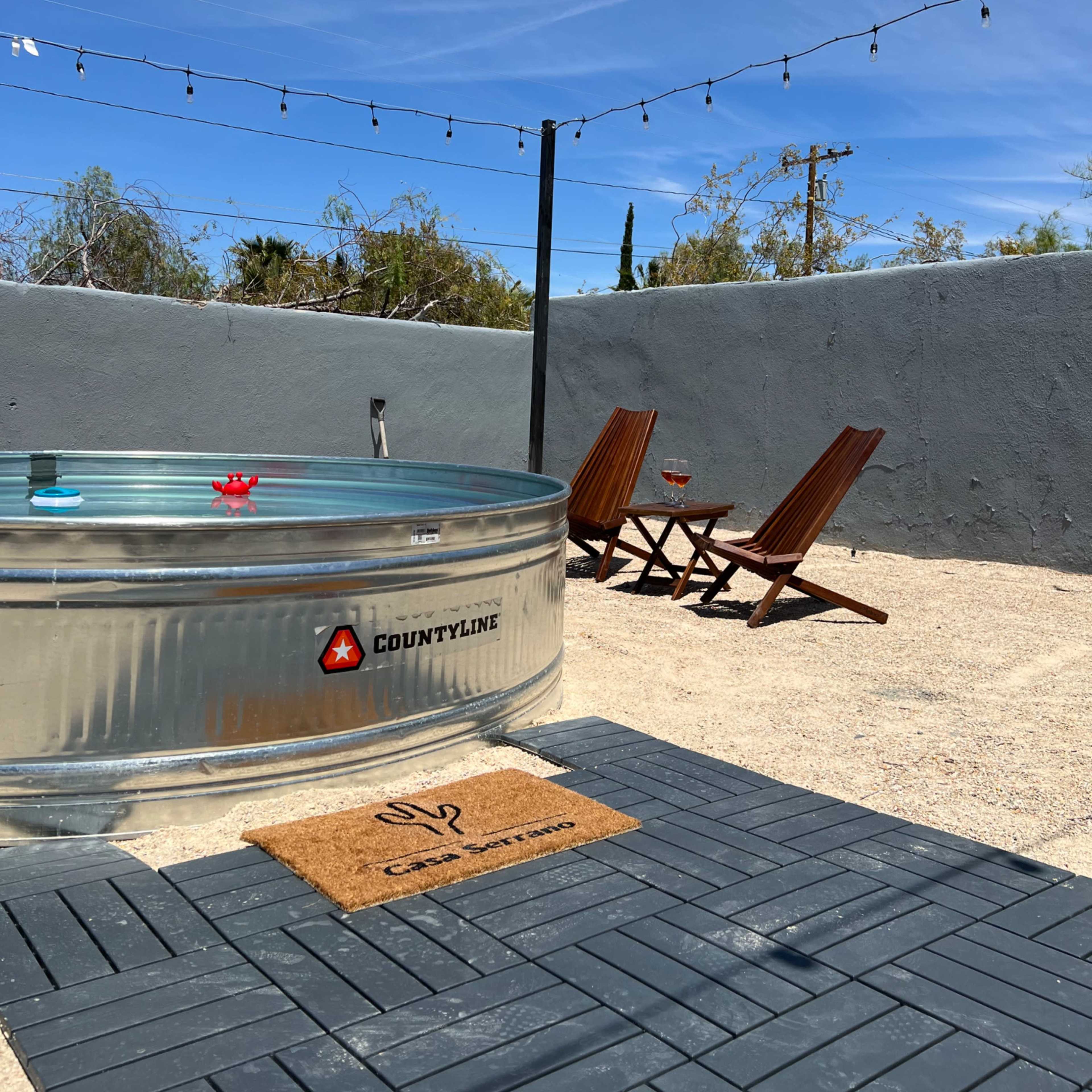 A metal hot tub sits on a gravel patio beside two wooden lounge chairs under string lights.
