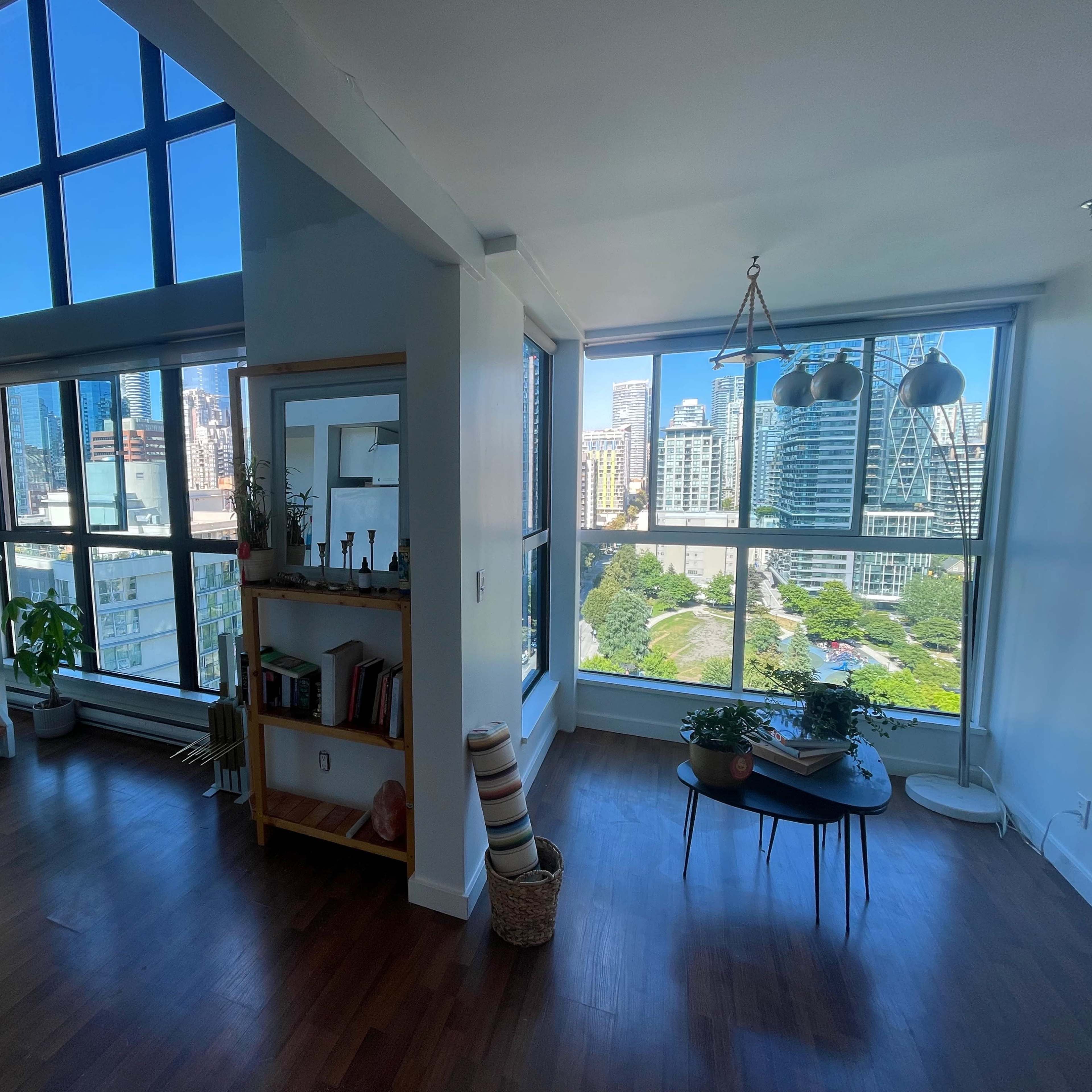 A bright, spacious living area features large windows with city views, a staircase, a bookshelf, and a small round table.