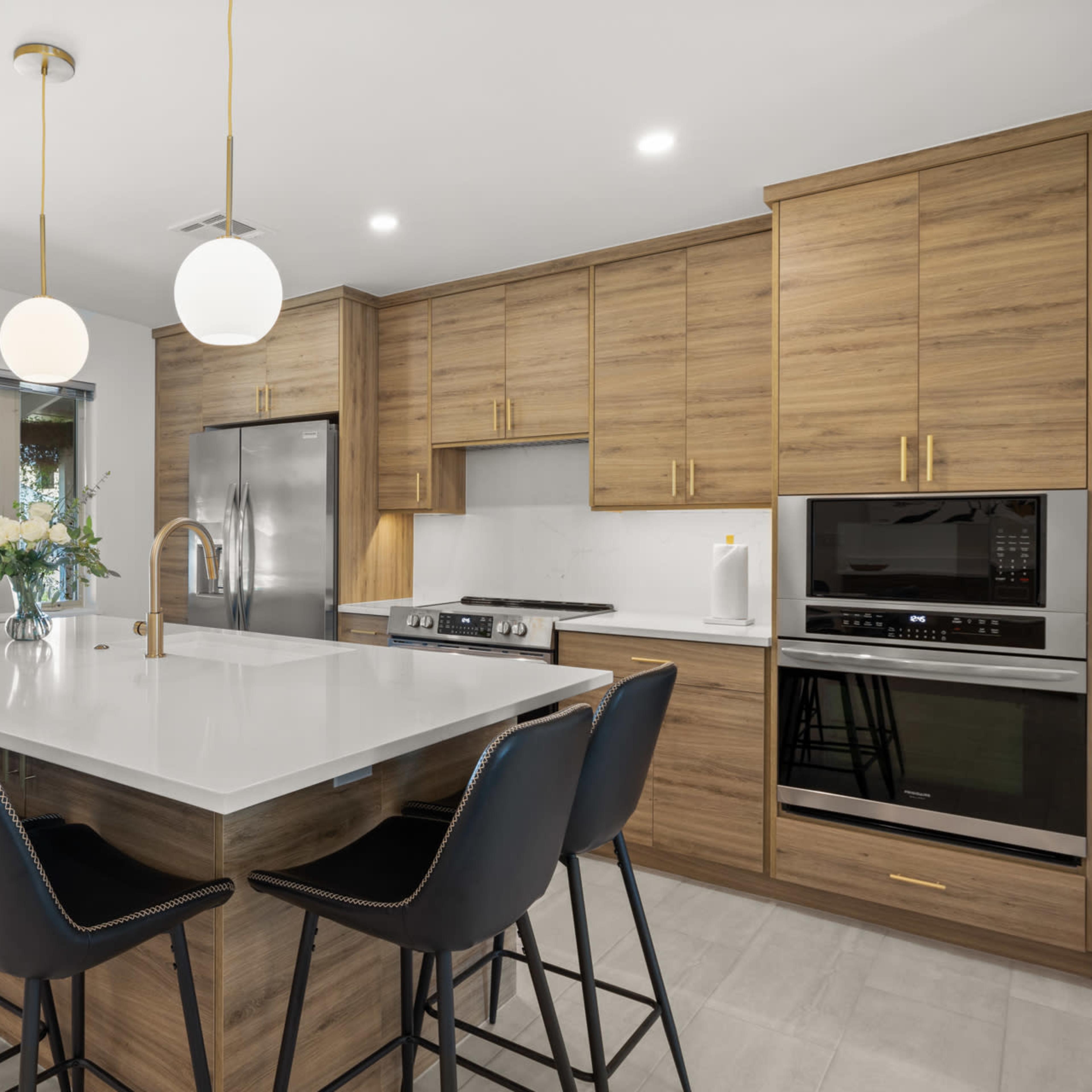 A modern kitchen features a large island with black chairs, wooden cabinetry, and pendant lights above.