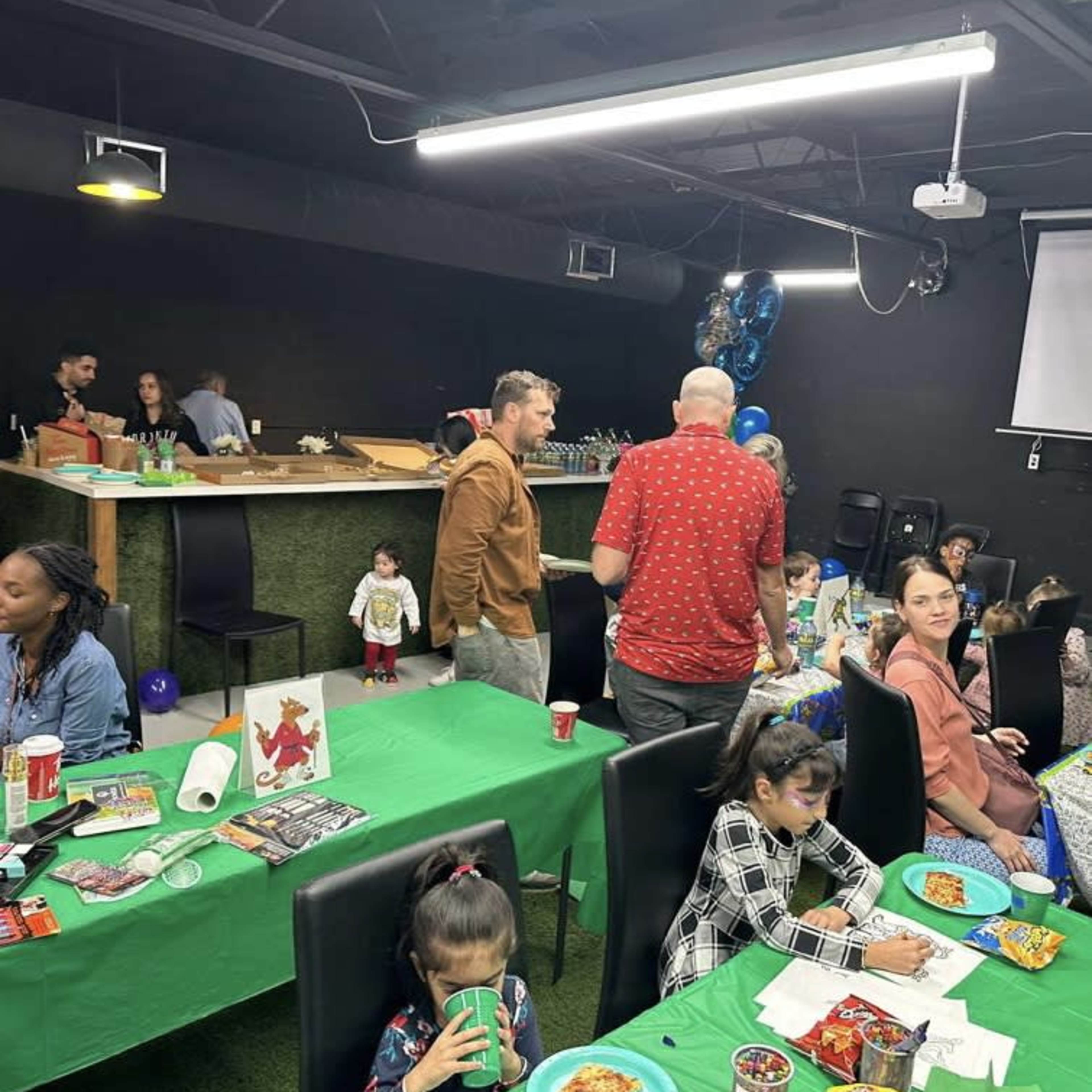A party is taking place in a room with green tables, where children are eating and playing while adults socialize in the background.
