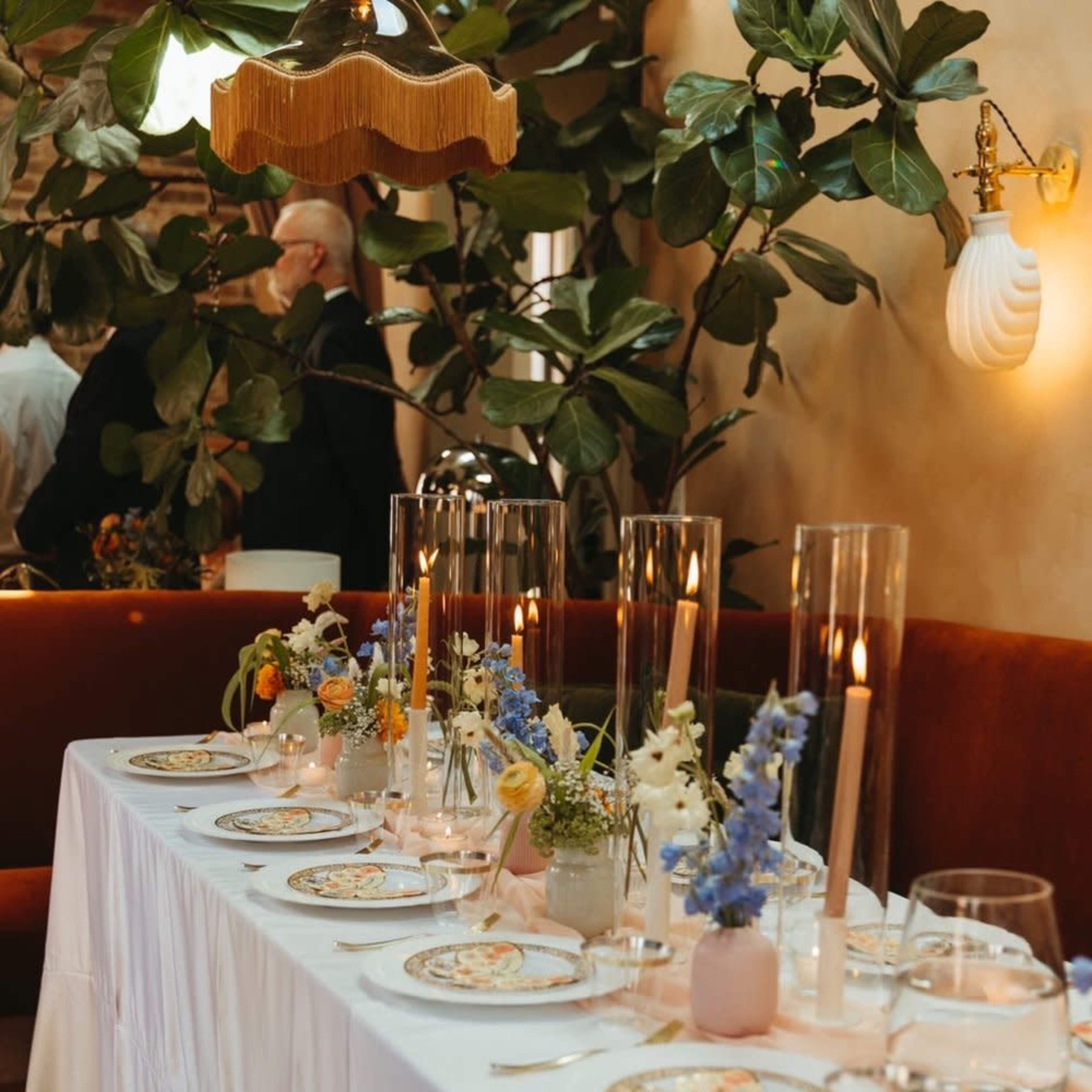 A long dining table is elegantly set with floral centerpieces, candles, and fine china, surrounded by greenery and soft lighting.