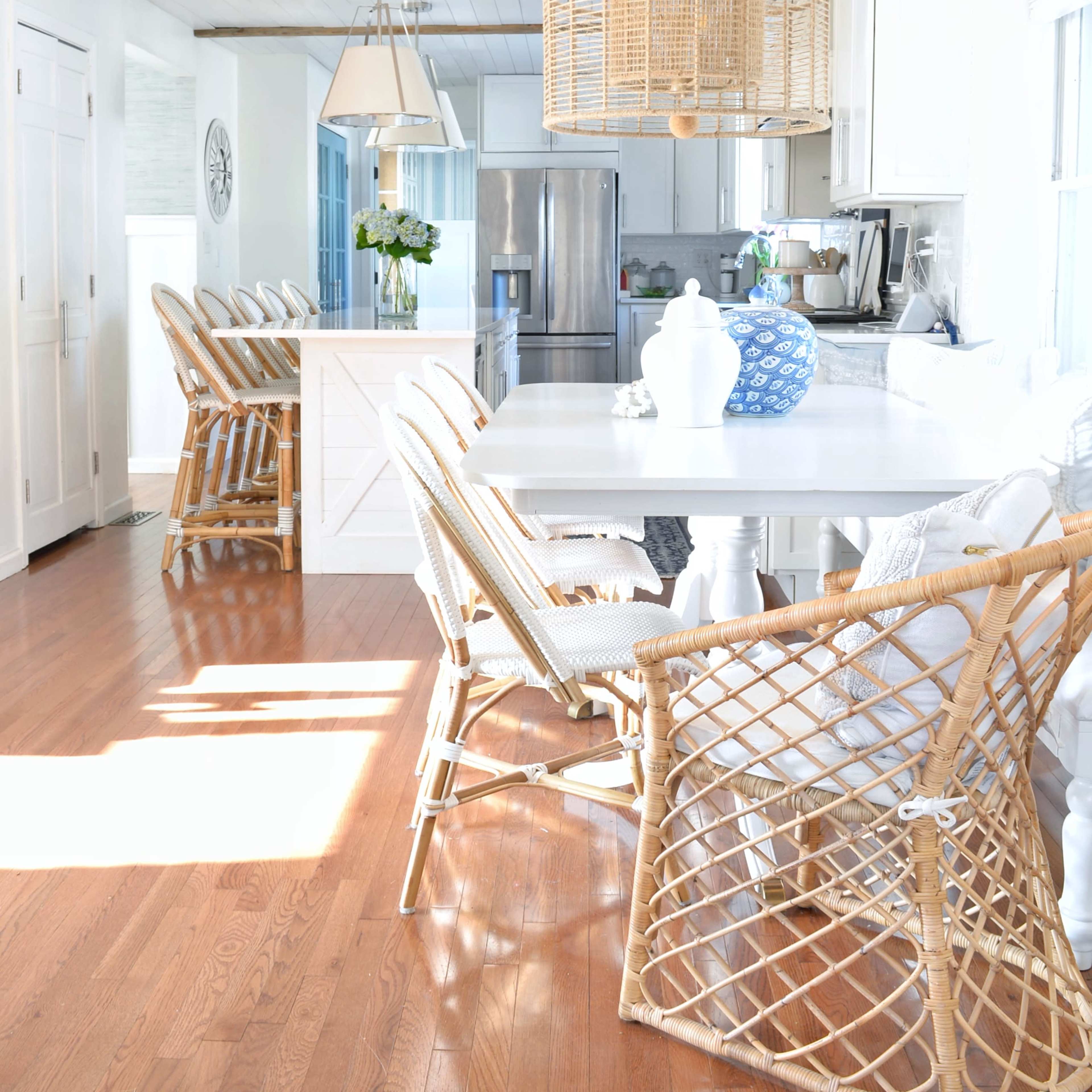 A bright, modern kitchen features a dining area with wooden flooring, a white table, and several wicker chairs arranged by the side.