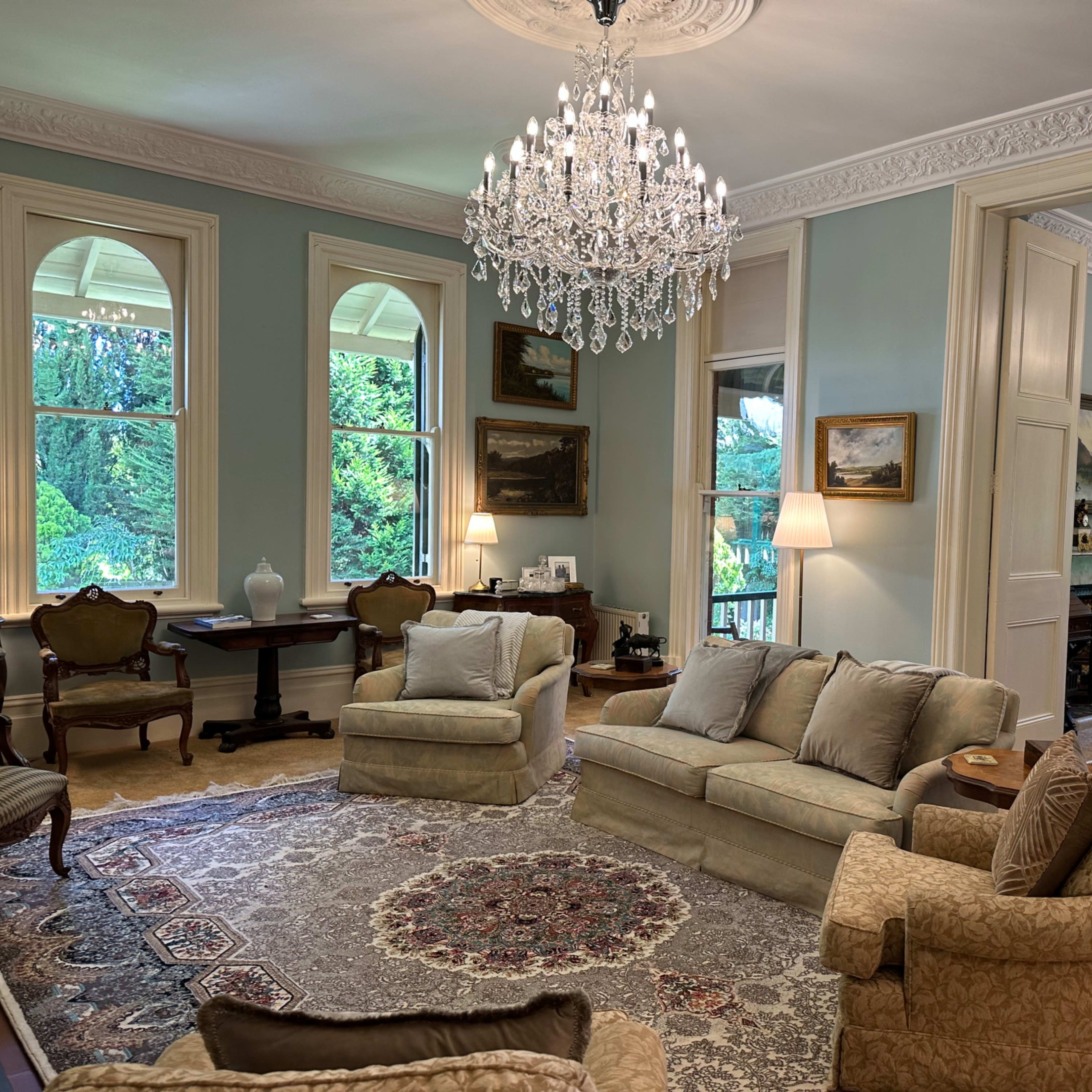 The room features two elegant chandeliers, a light blue color scheme, antique furniture, and large windows overlooking a garden.