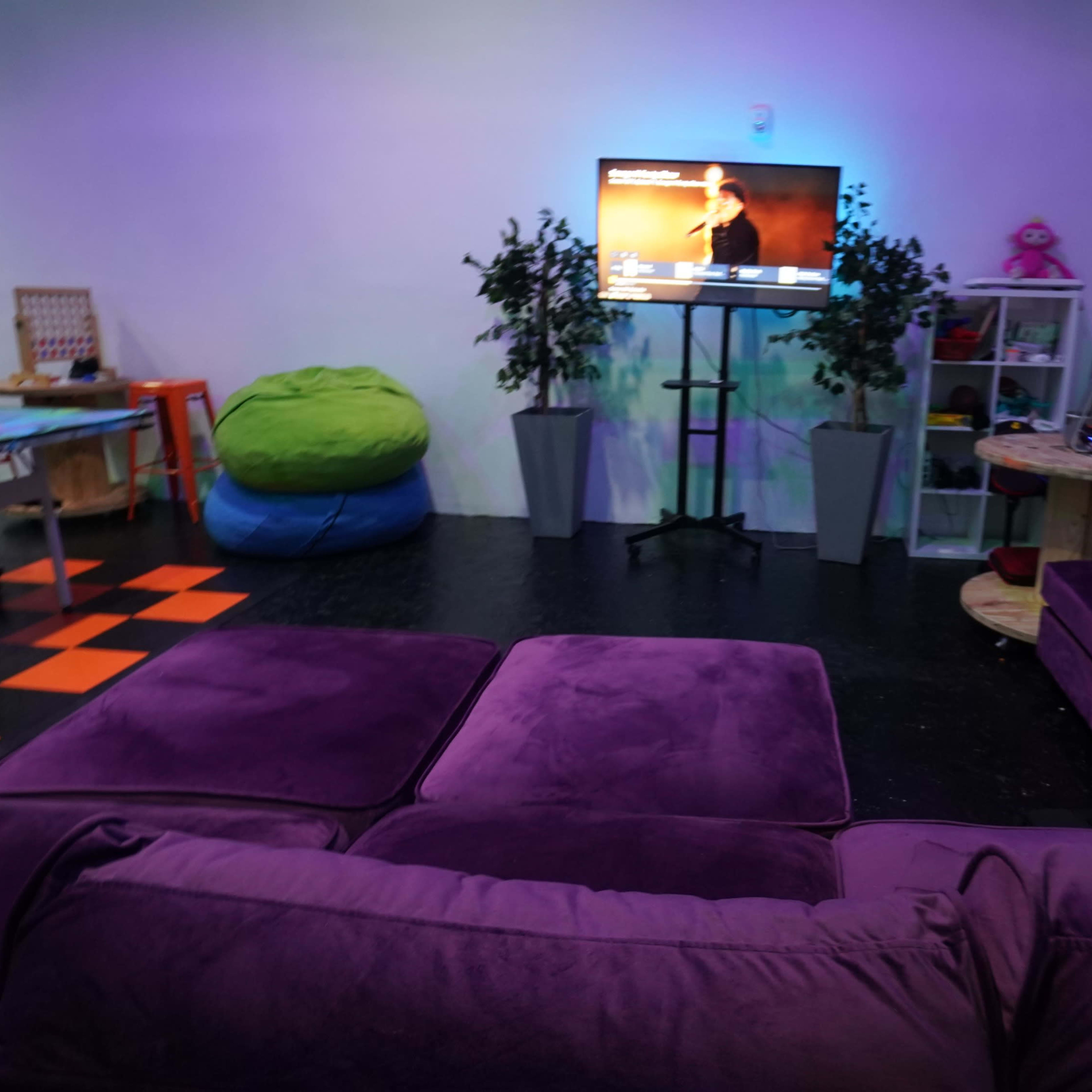 A cozy indoor space featuring a purple sofa, green bean bags, a television on a stand, and decorative plants.