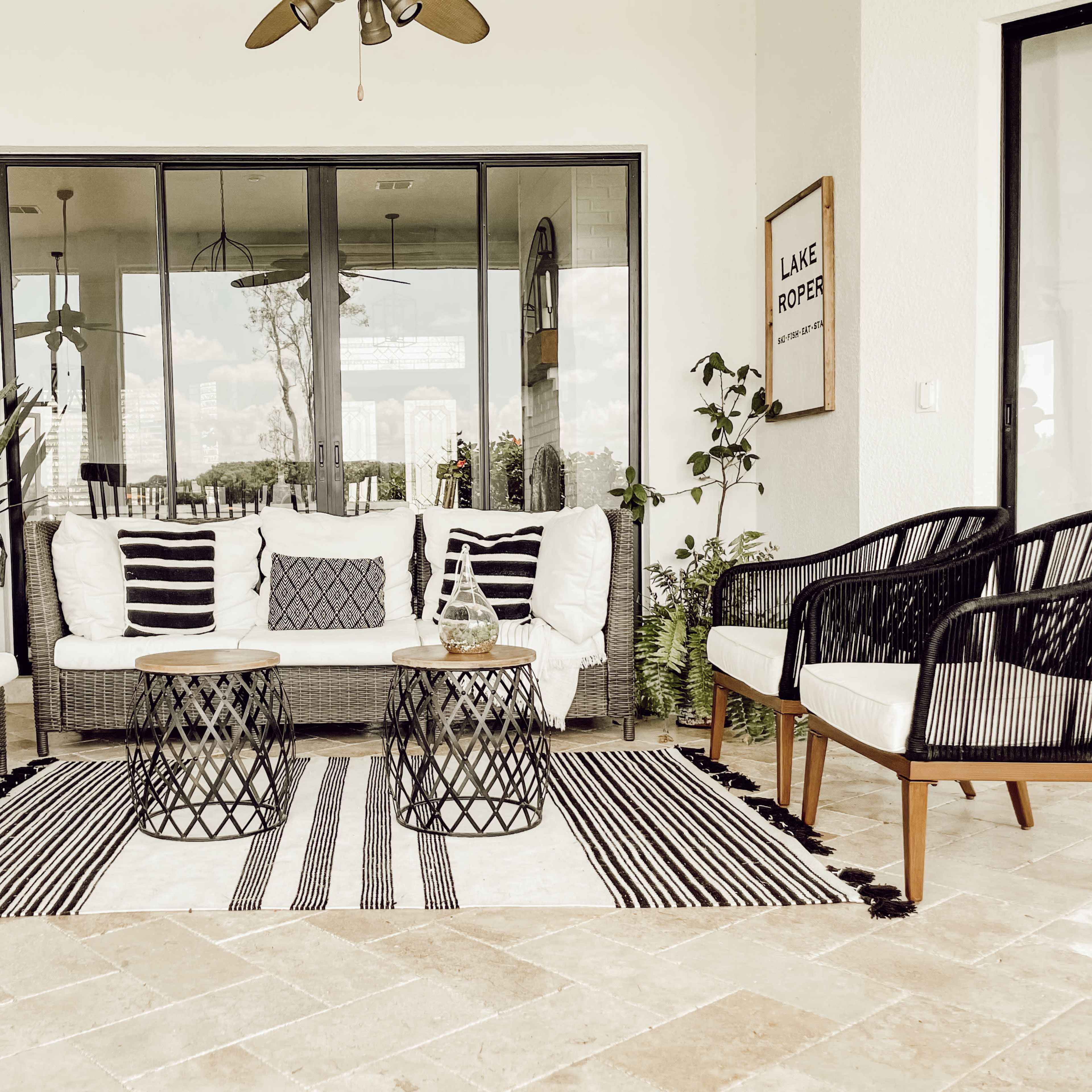 A cozy outdoor seating area with a white sofa, two black chairs, a patterned rug, and two round tables surrounded by plants.