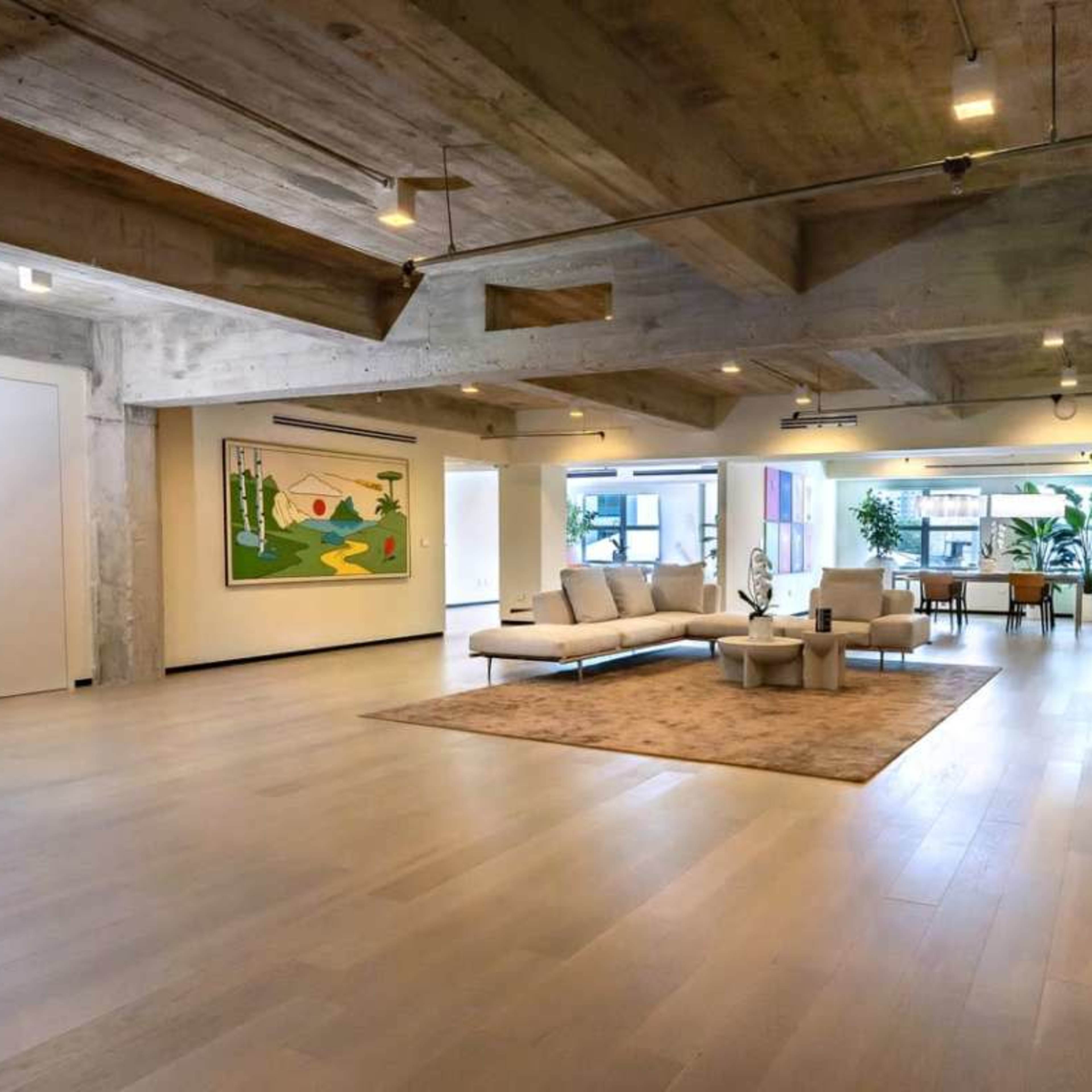A spacious, modern living area with a large sectional sofa, a coffee table, and decorative plants, complemented by exposed ceiling beams and large windows.