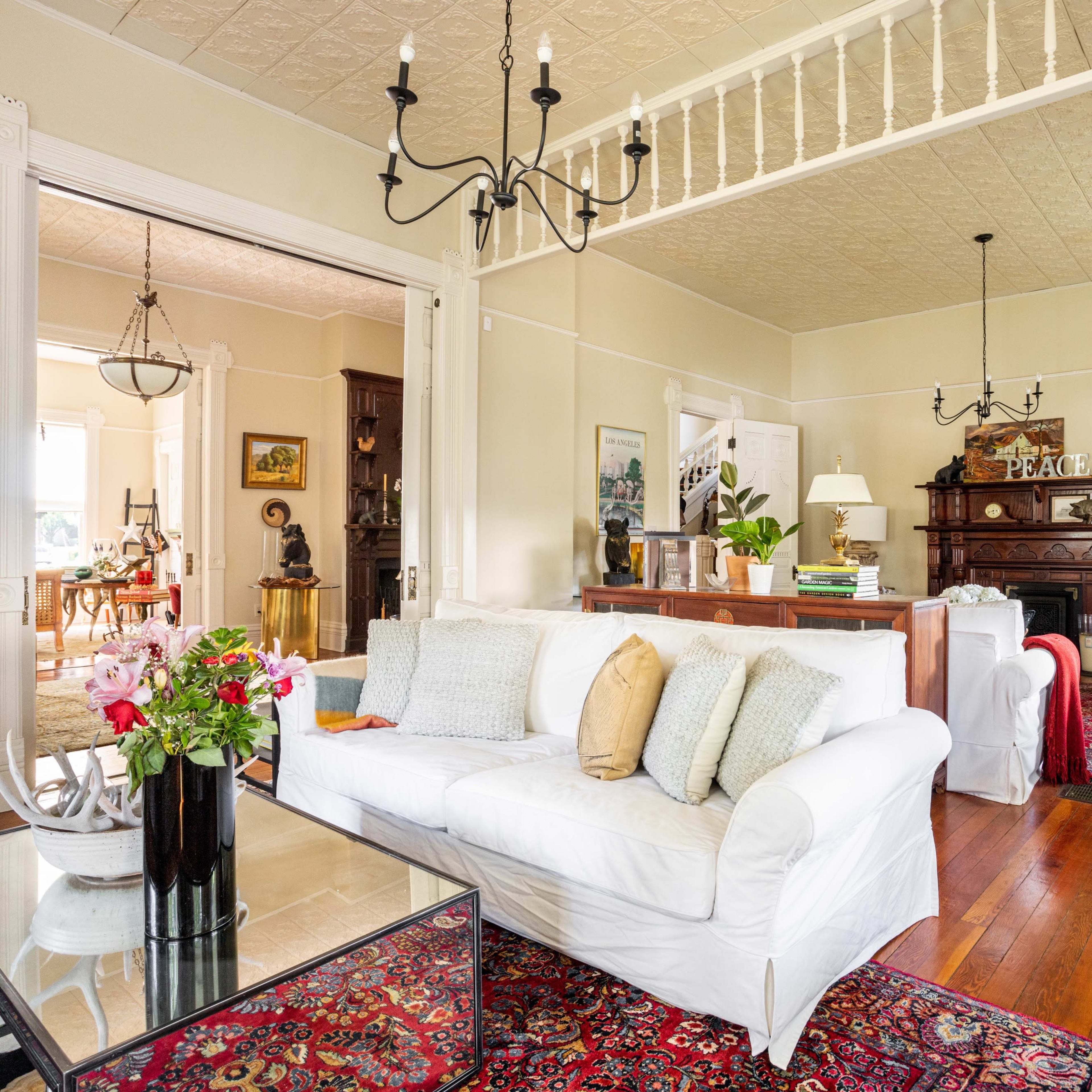 A spacious living room with white furniture, hardwood floors, and ornate decorations, leading to another room visible in the background.