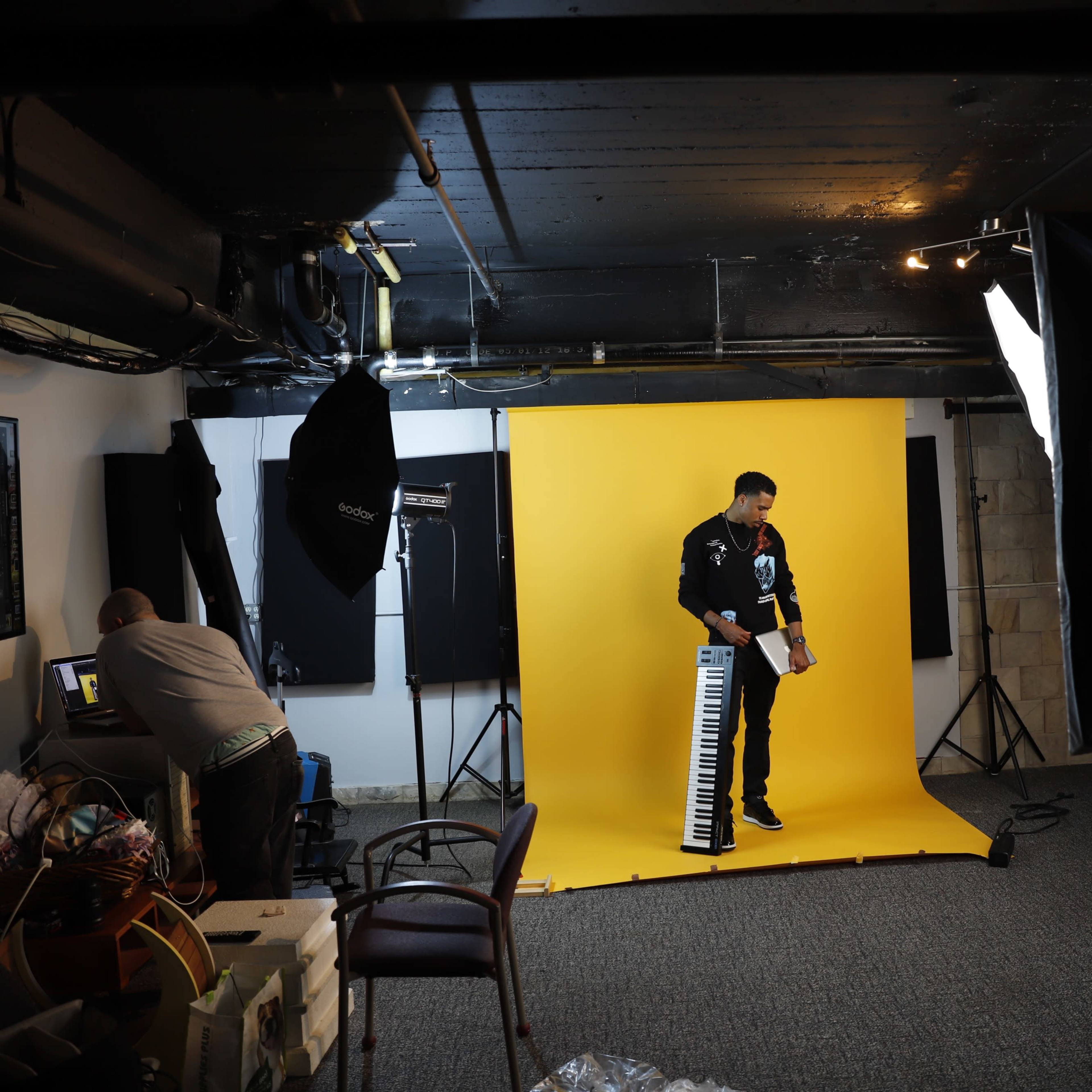 A musician stands with a keyboard in front of a yellow backdrop while a photographer edits images on a computer in a studio setting.