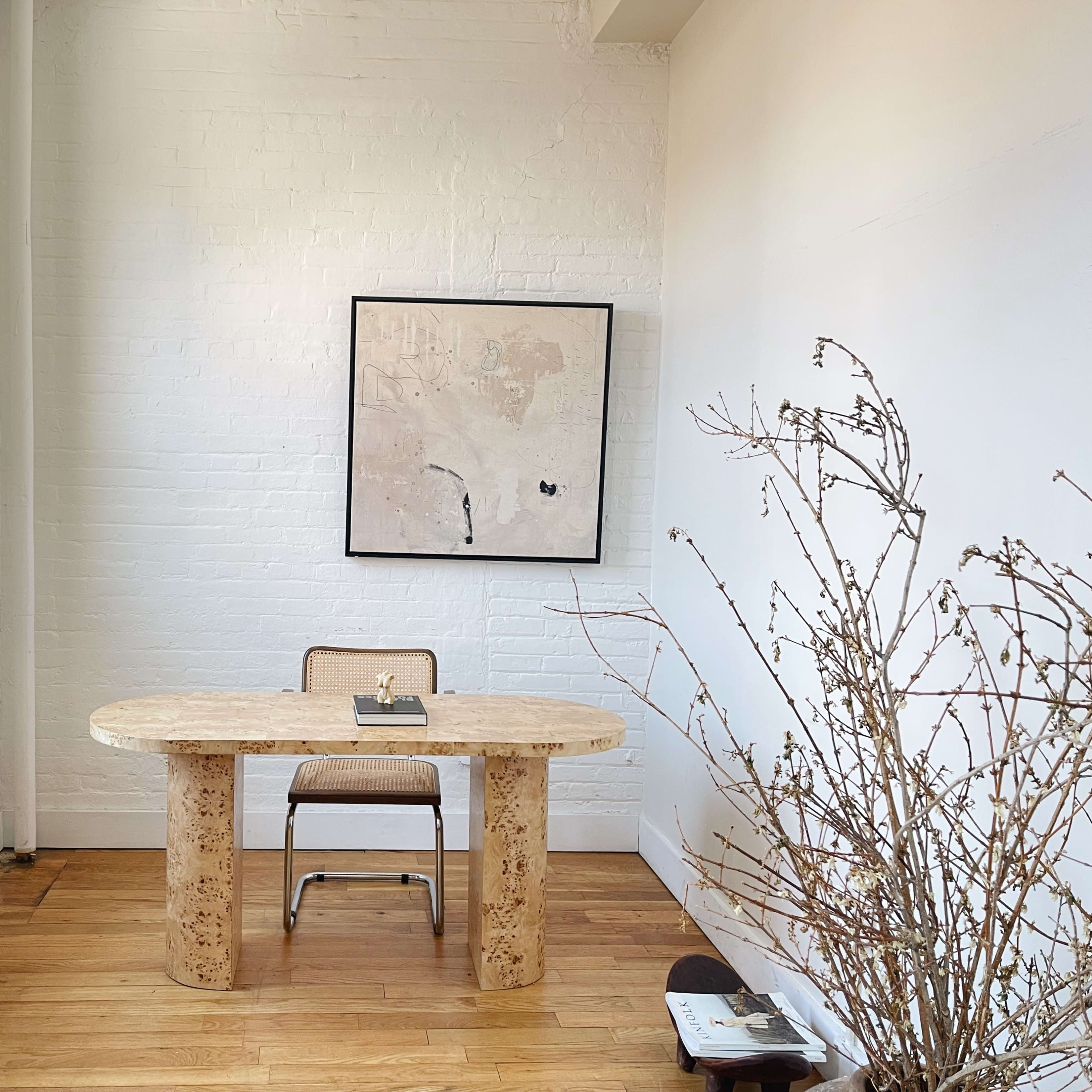 A minimalist workspace featuring a round, stone table with a chair, a small lamp, an artwork on the wall, and a decorative branch in a corner.
