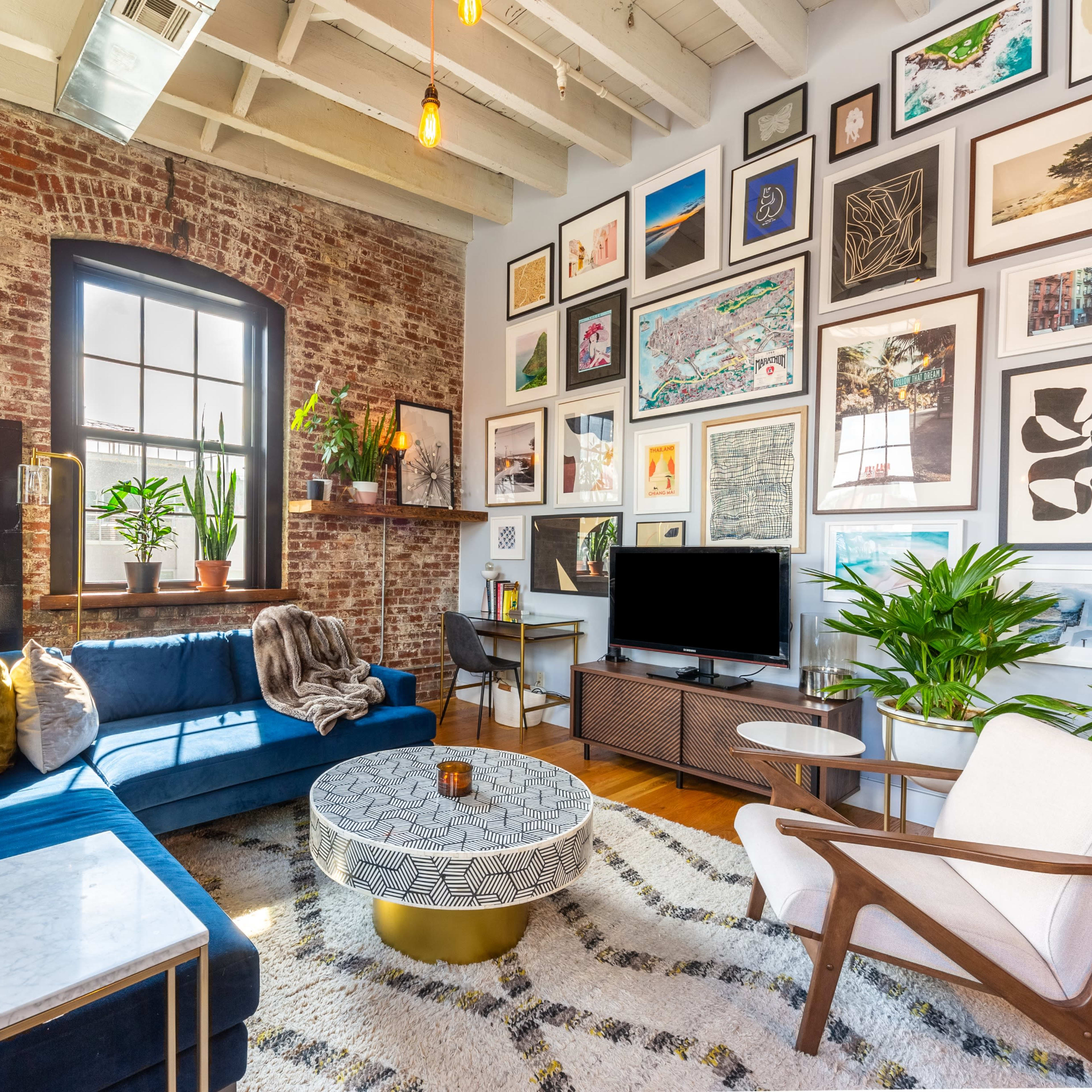 The scene shows a stylish living room with exposed brick walls, a blue sofa, a round coffee table, and a gallery wall filled with various framed artworks.