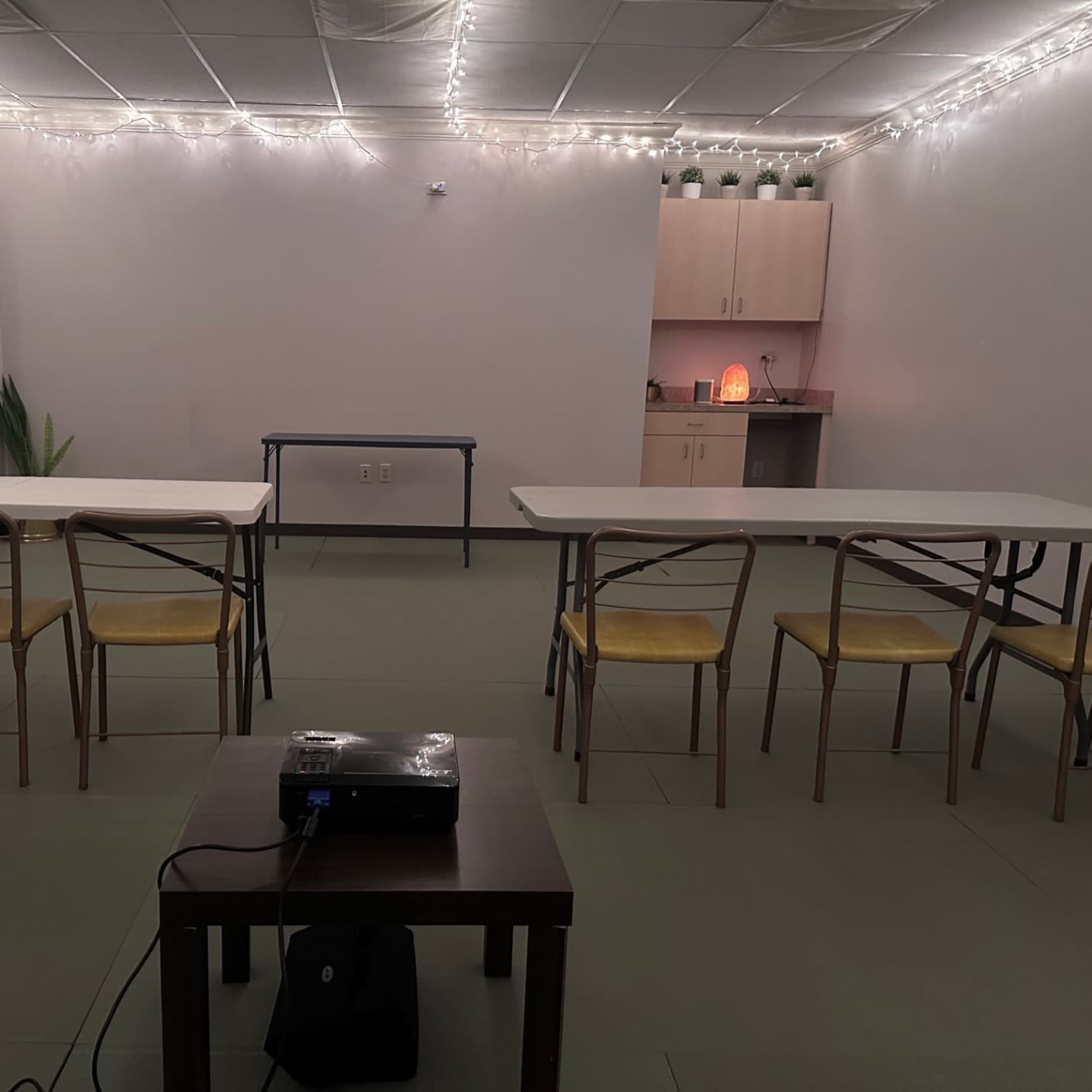 A classroom setting features two tables arranged with chairs on either side, a projector on a coffee table, and softly lit ambient lighting in the background.