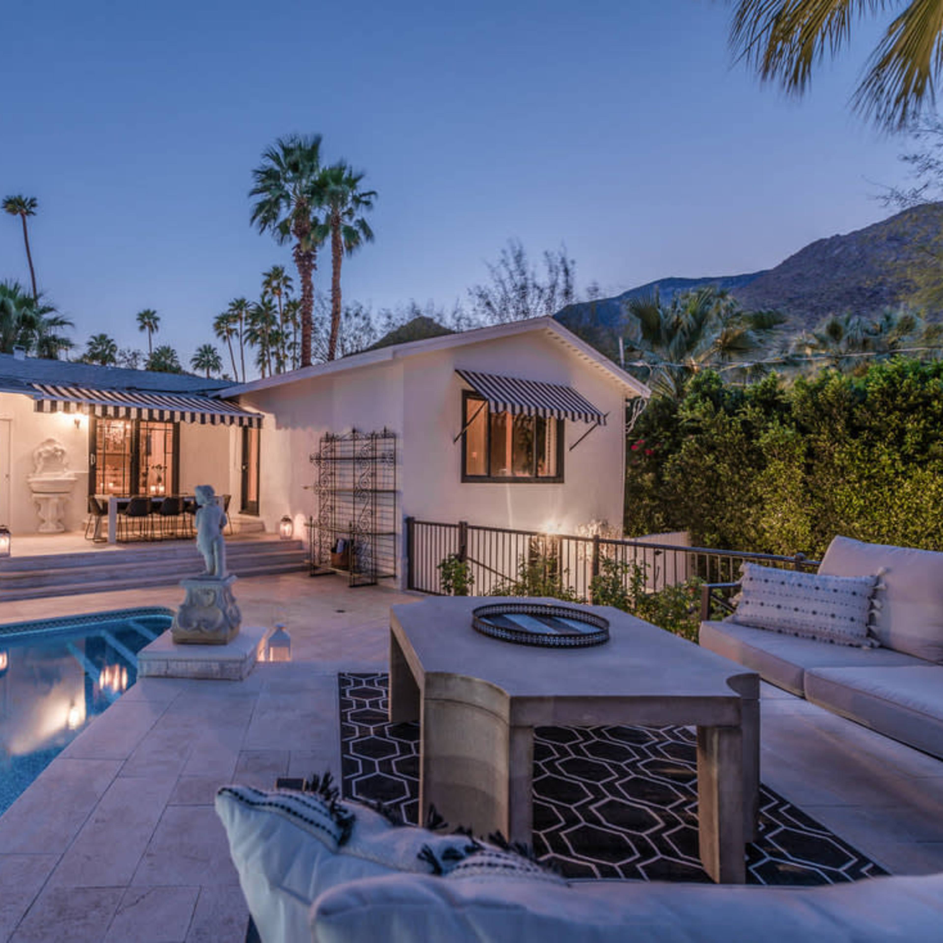 A serene outdoor space featuring a swimming pool, a table, and lounge seating, surrounded by palm trees and mountains under a twilight sky.