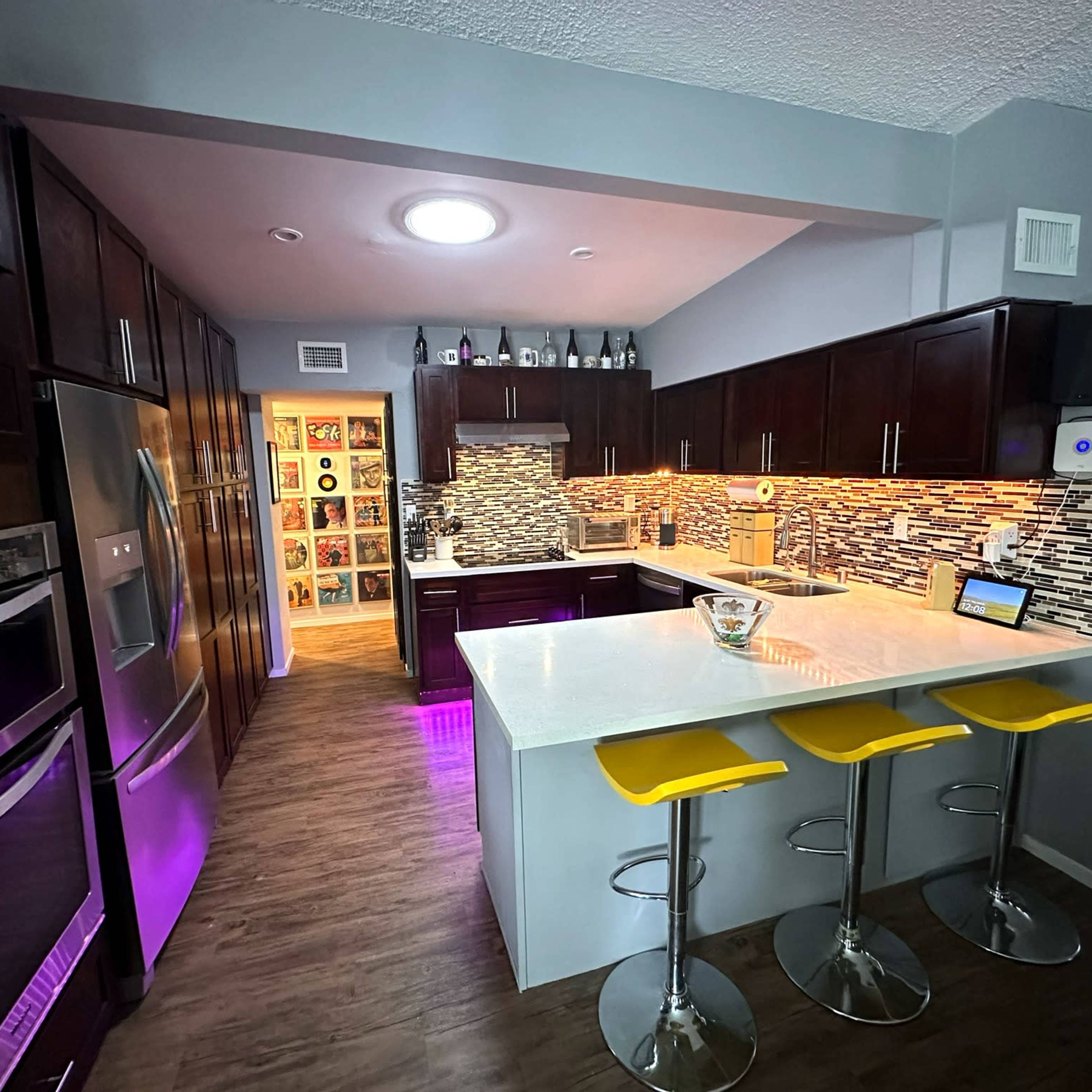 A modern kitchen featuring dark wood cabinets, stainless steel appliances, a white countertop, and yellow bar stools.