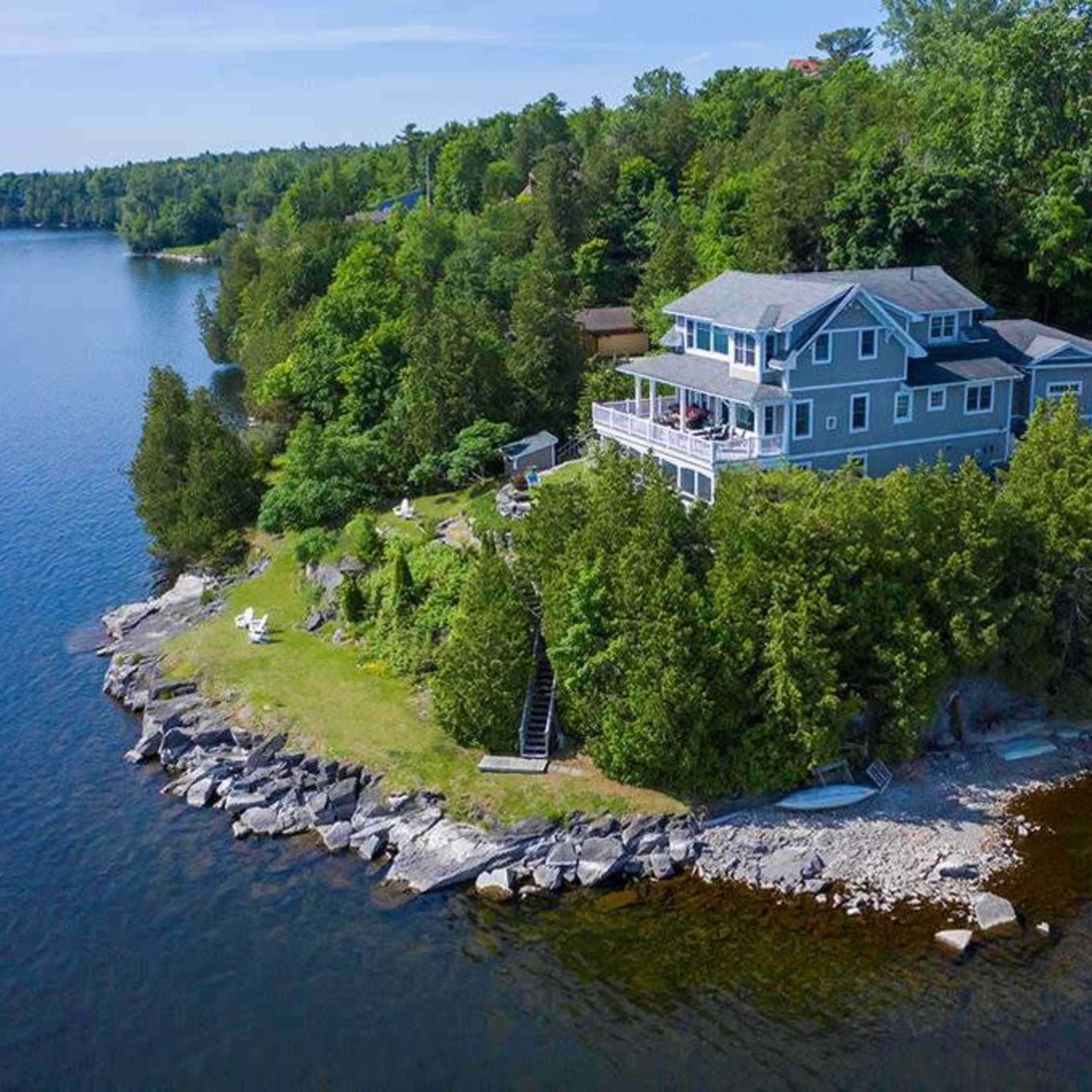 A blue house is situated on a rocky peninsula surrounded by water and dense greenery.