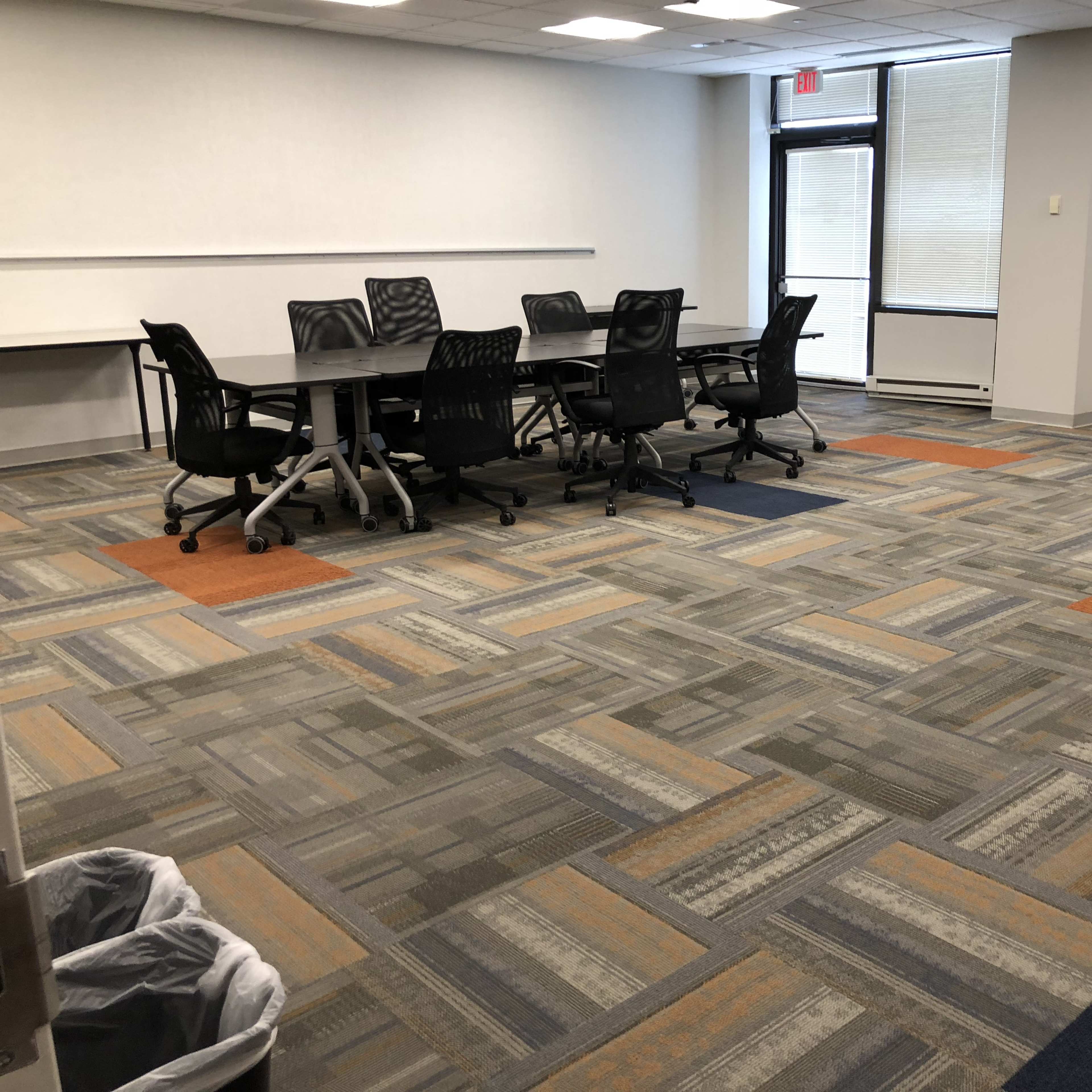 An empty conference room features a large table surrounded by black chairs on a patterned carpet with sections of orange and gray.