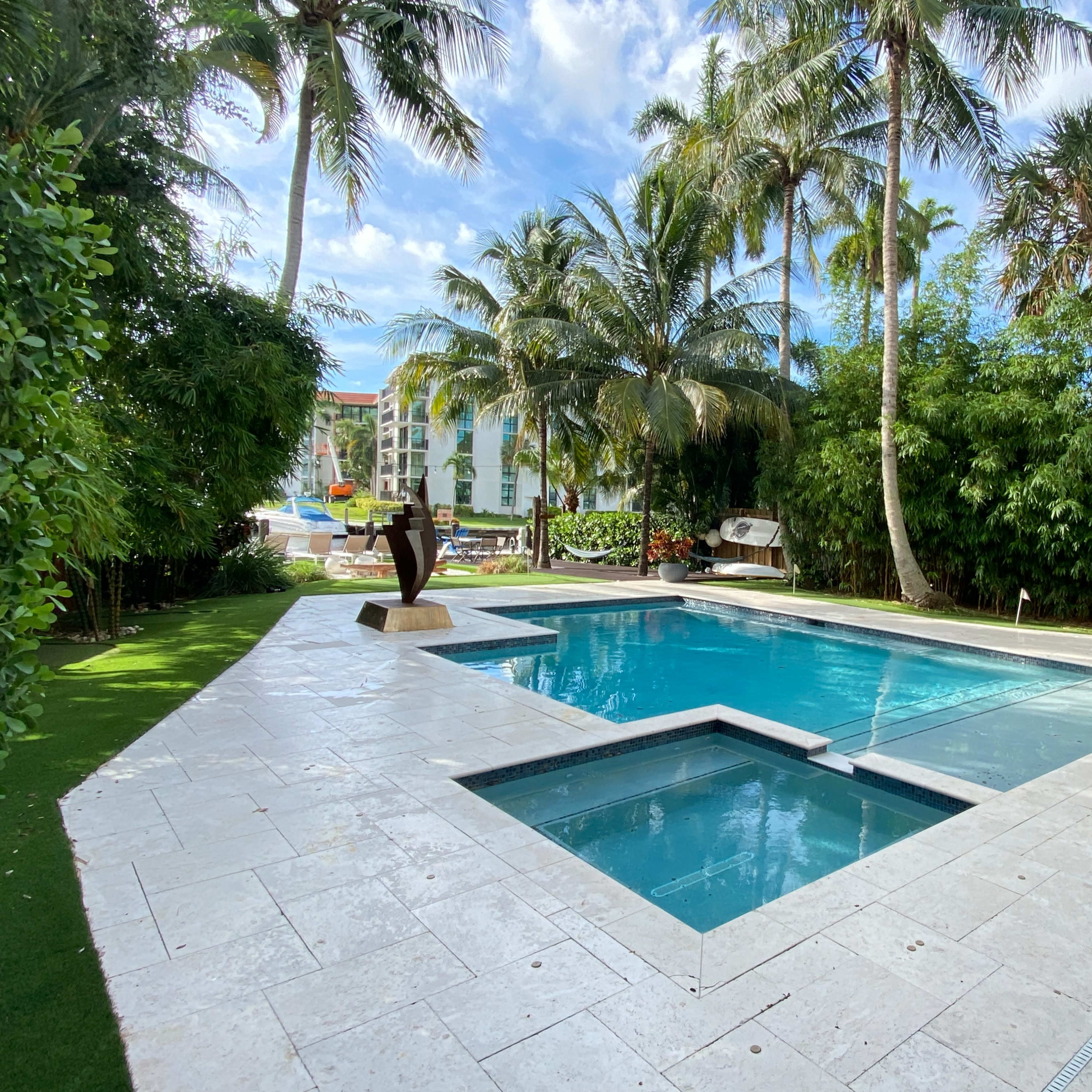 A landscaped backyard features a blue swimming pool with a spa, surrounded by palm trees and decorative foliage.