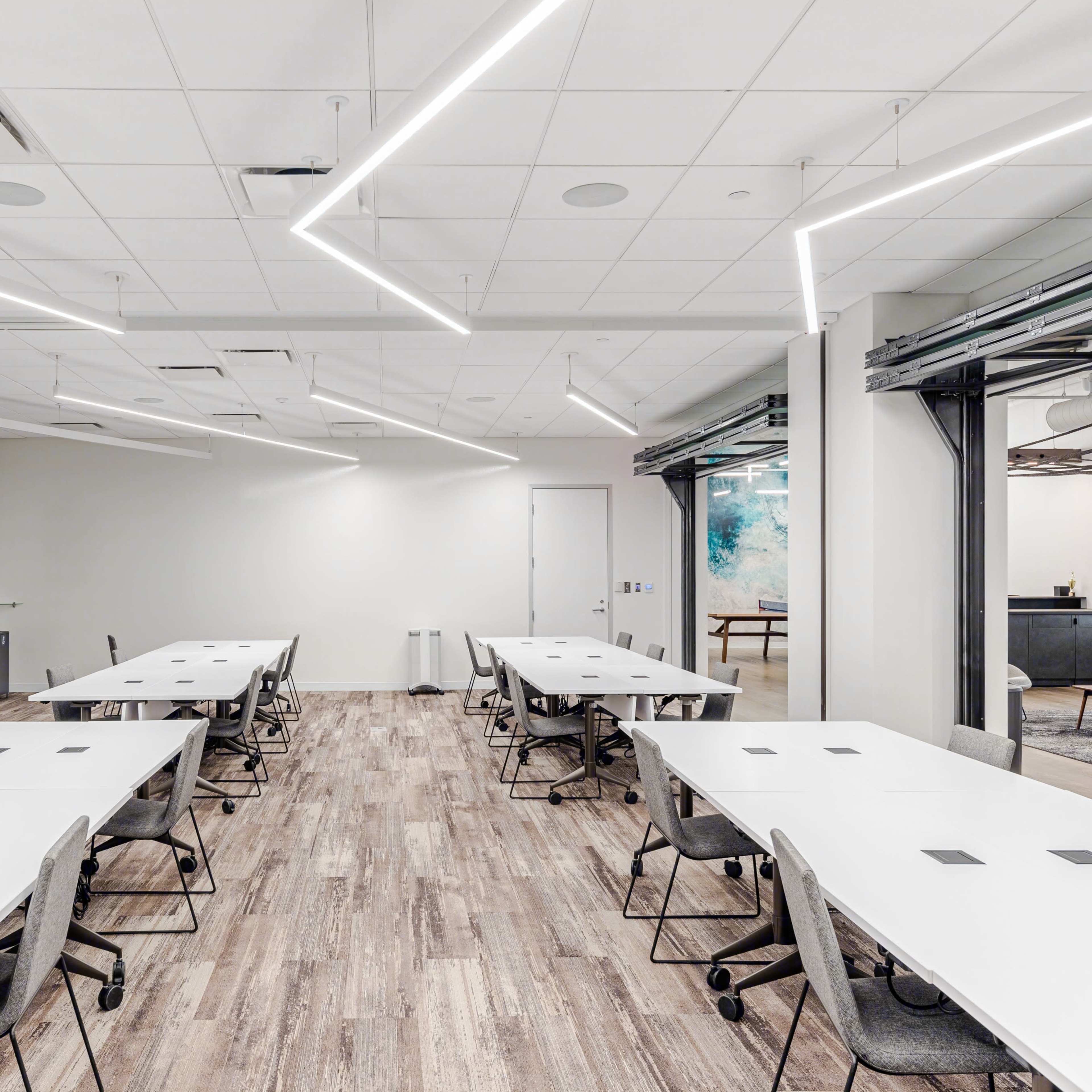 A modern conference room with multiple rectangular tables arranged in a collaborative layout, illuminated by angular ceiling lights.
