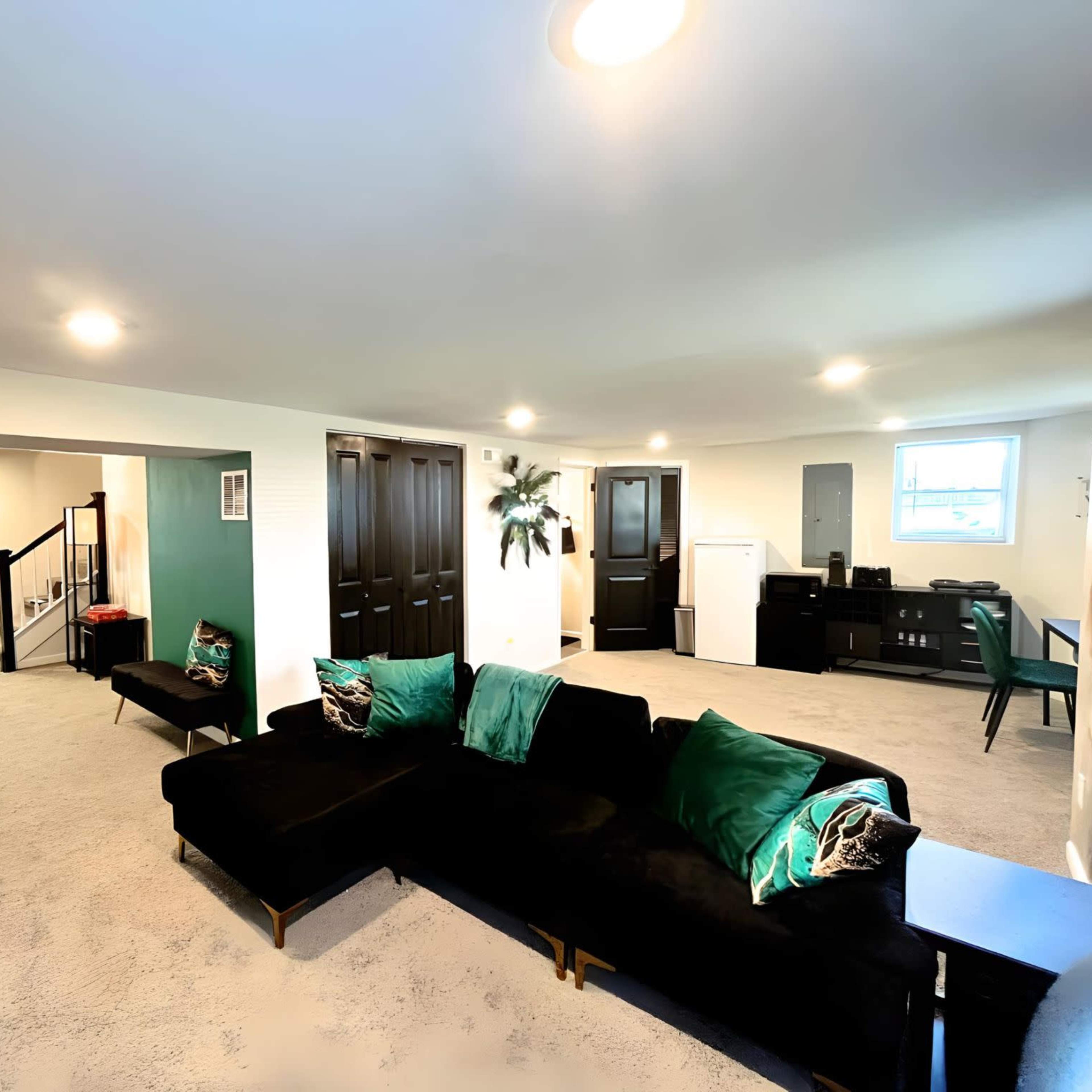 A spacious, carpeted basement with a black sectional sofa, a small desk, and multiple doors leading to separate rooms.