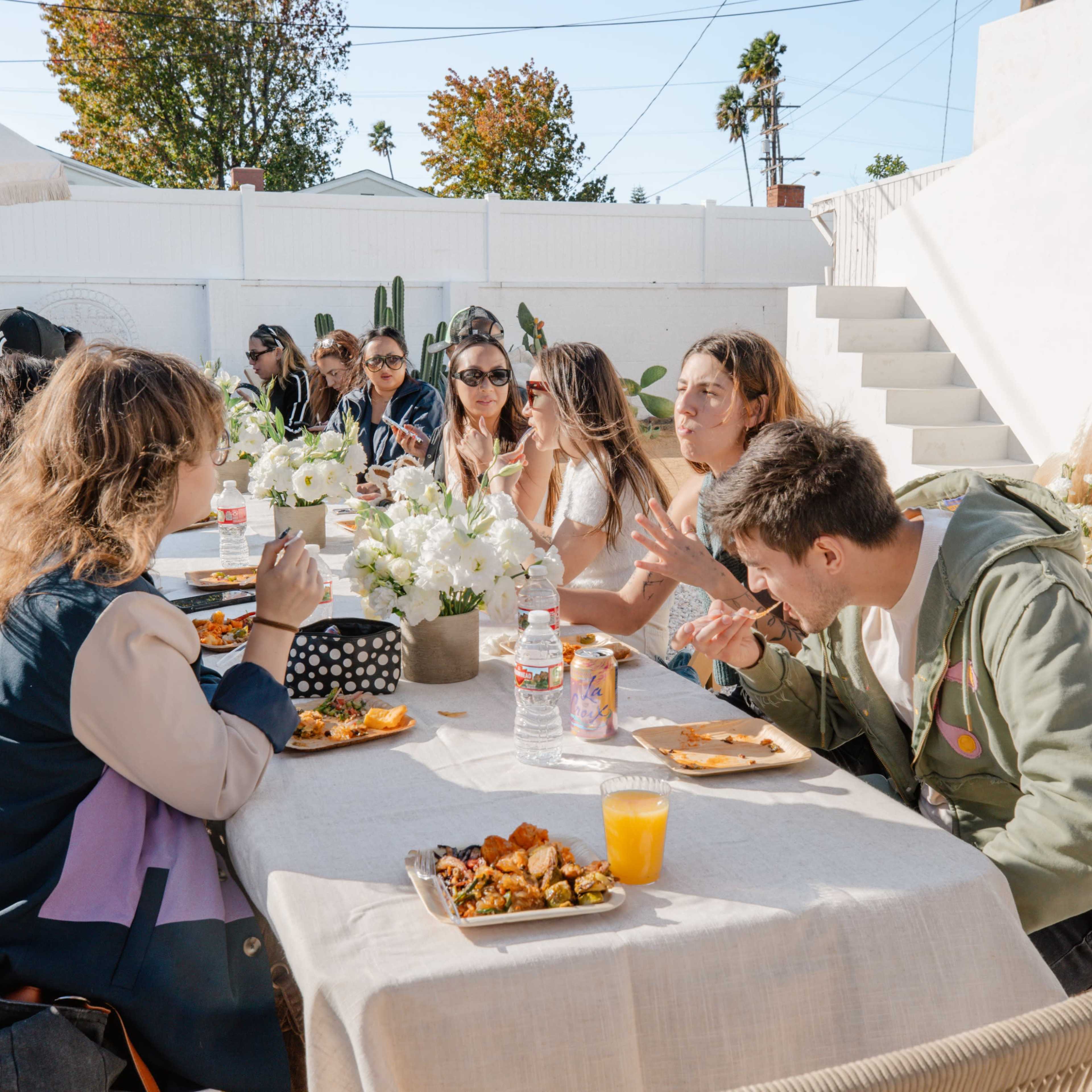 A group of people sits around a long table outdoors, enjoying food and conversation in a sunlit setting.