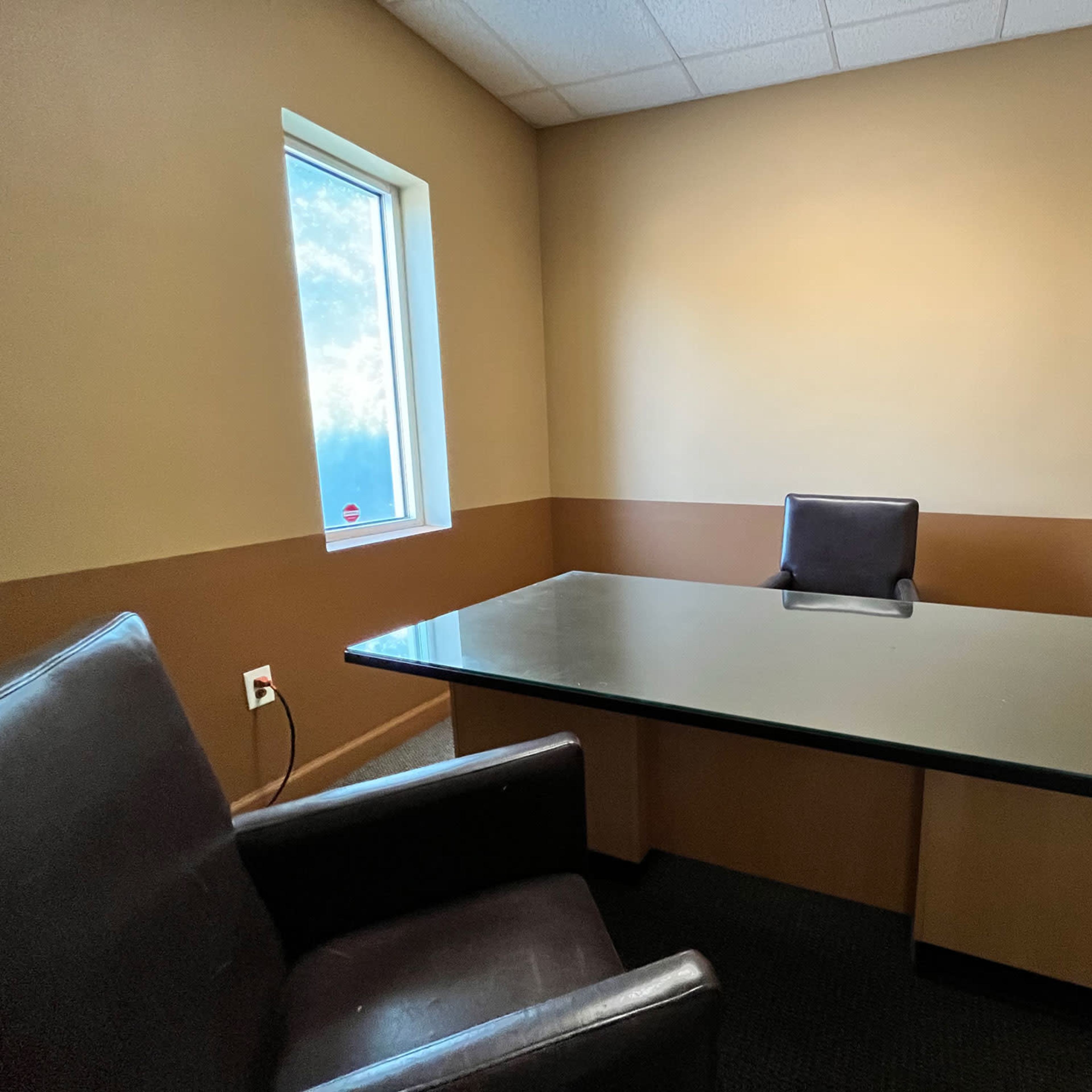 An empty office space with a glass-top desk and a single black chair facing a window, alongside two brown chairs.