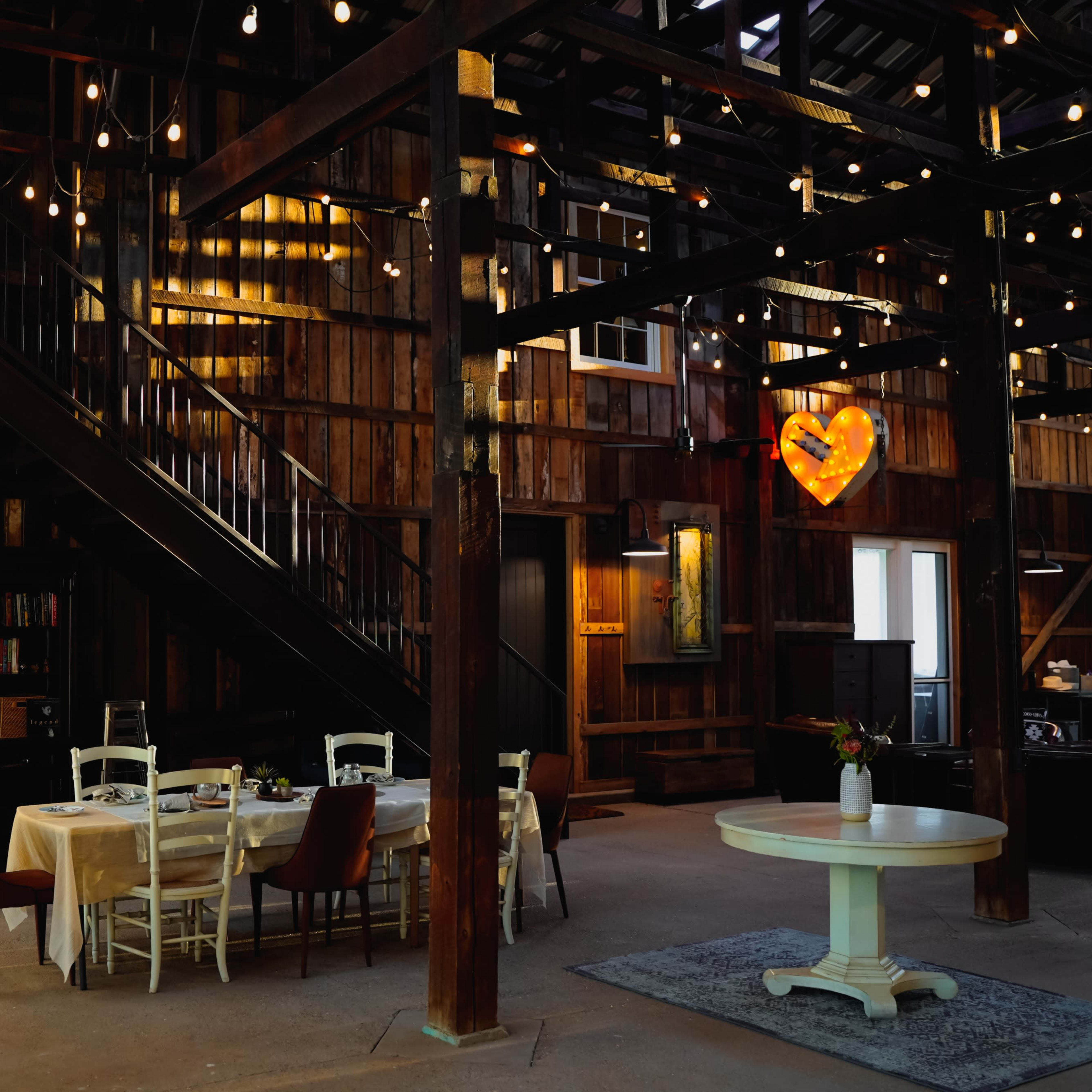 The interior of a rustic, wooden barn features a dining area with a table and chairs beneath string lights and a heart-shaped decor piece.