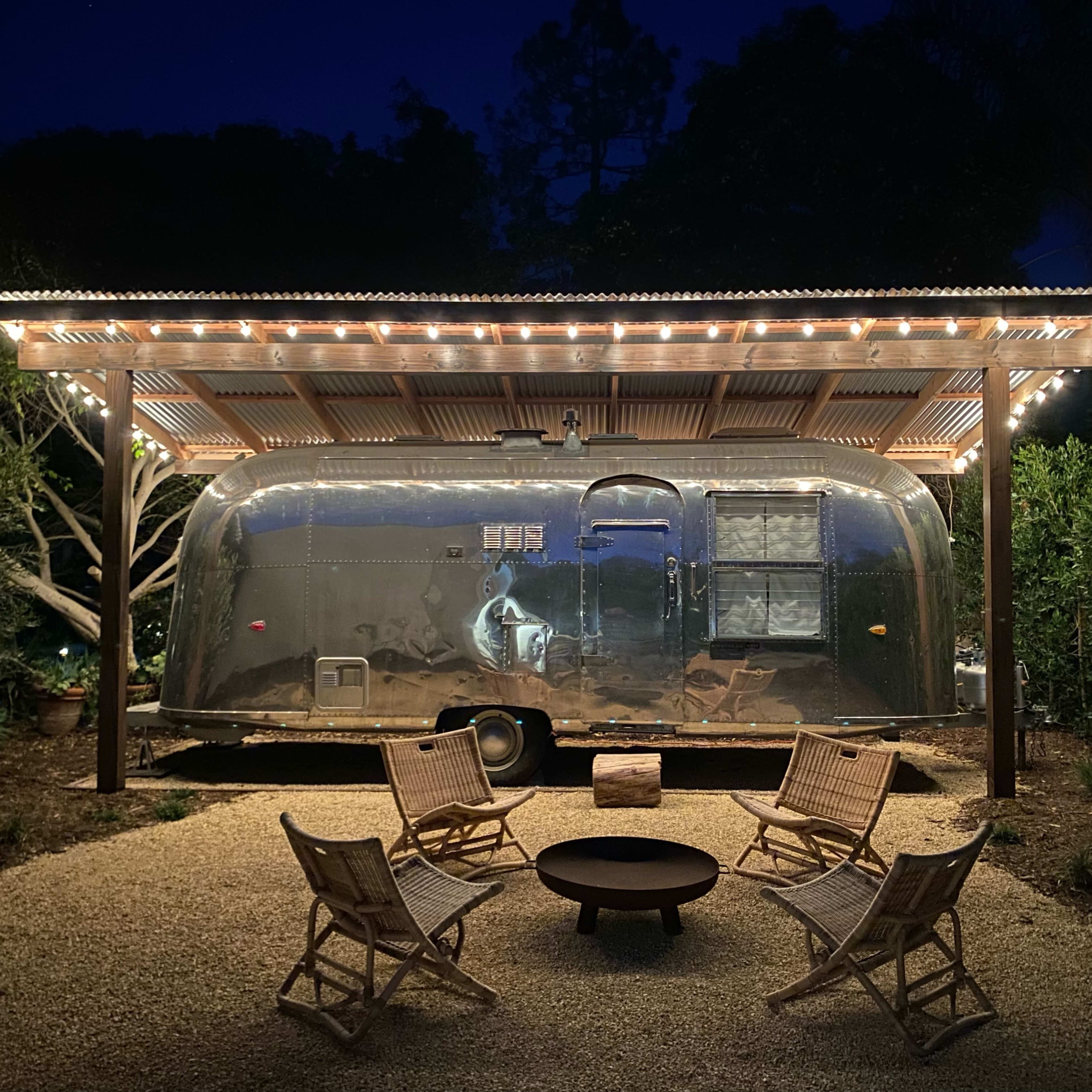 An Airstream trailer is parked under a lit wooden pergola surrounded by chairs and a fire pit at night.