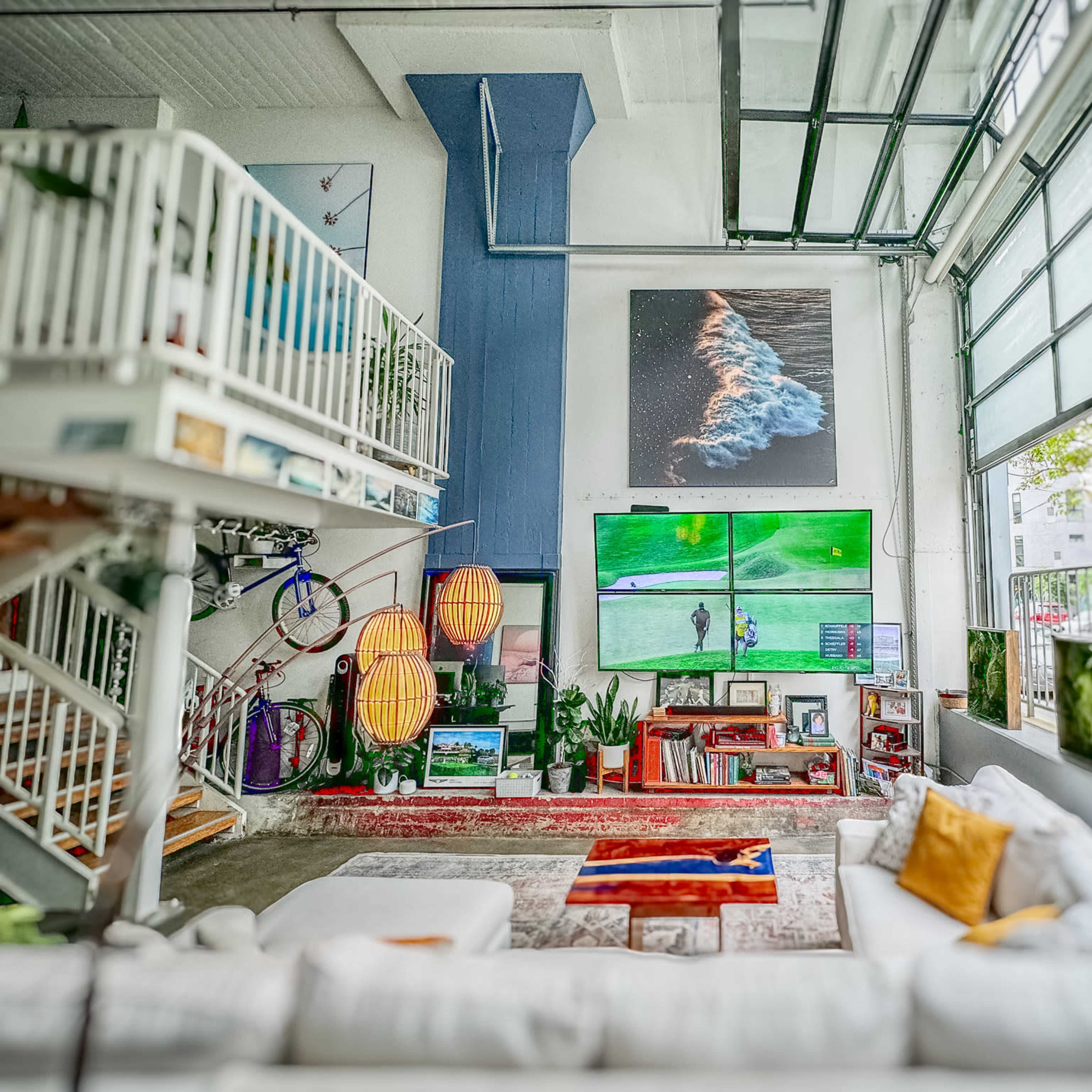 A spacious living area featuring a large sectional sofa, wall-mounted televisions displaying a golf game, and decorative items arranged around an open layout with industrial-style architecture.