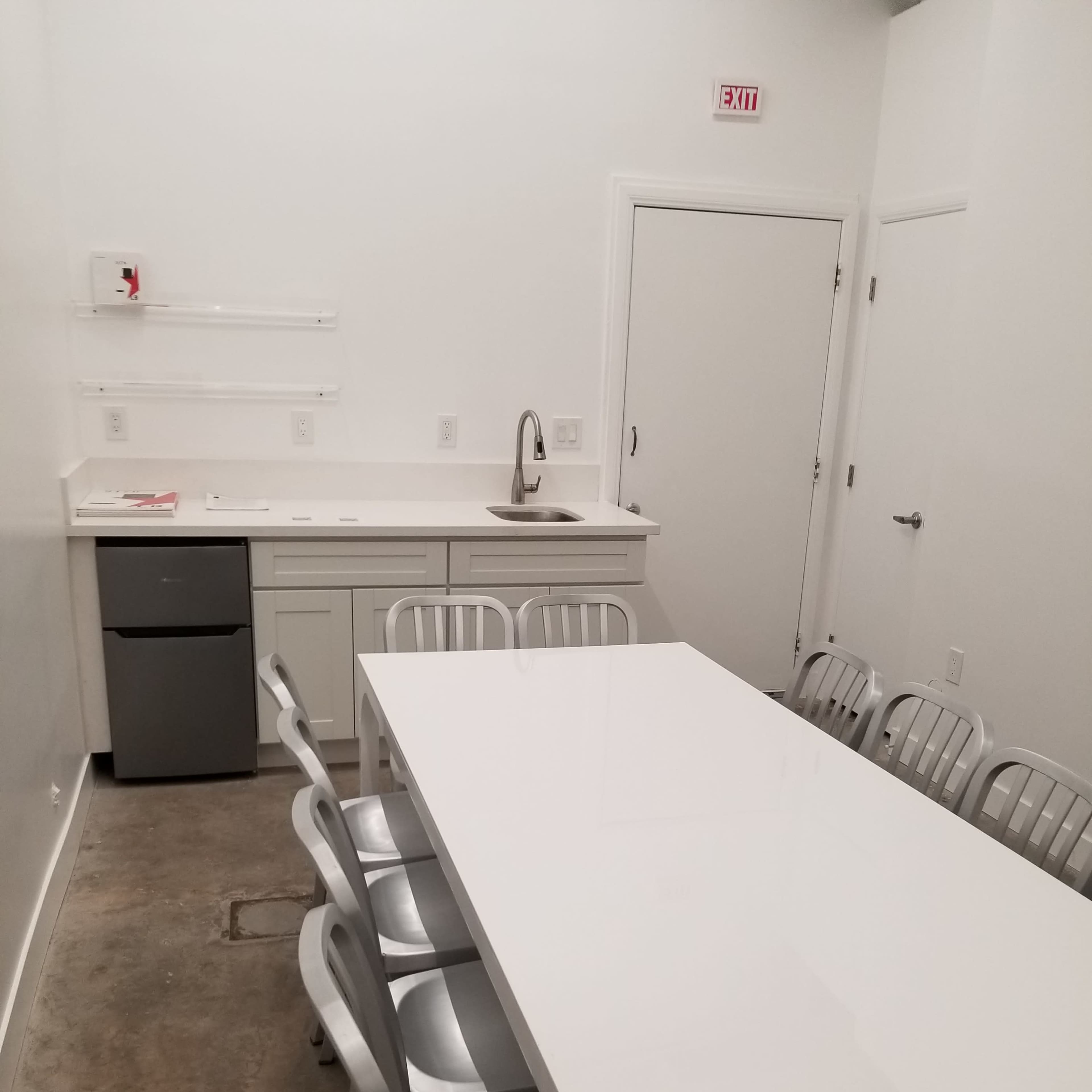 A modern kitchen with a long white table and metal chairs, a sink, and a small refrigerator against the wall.
