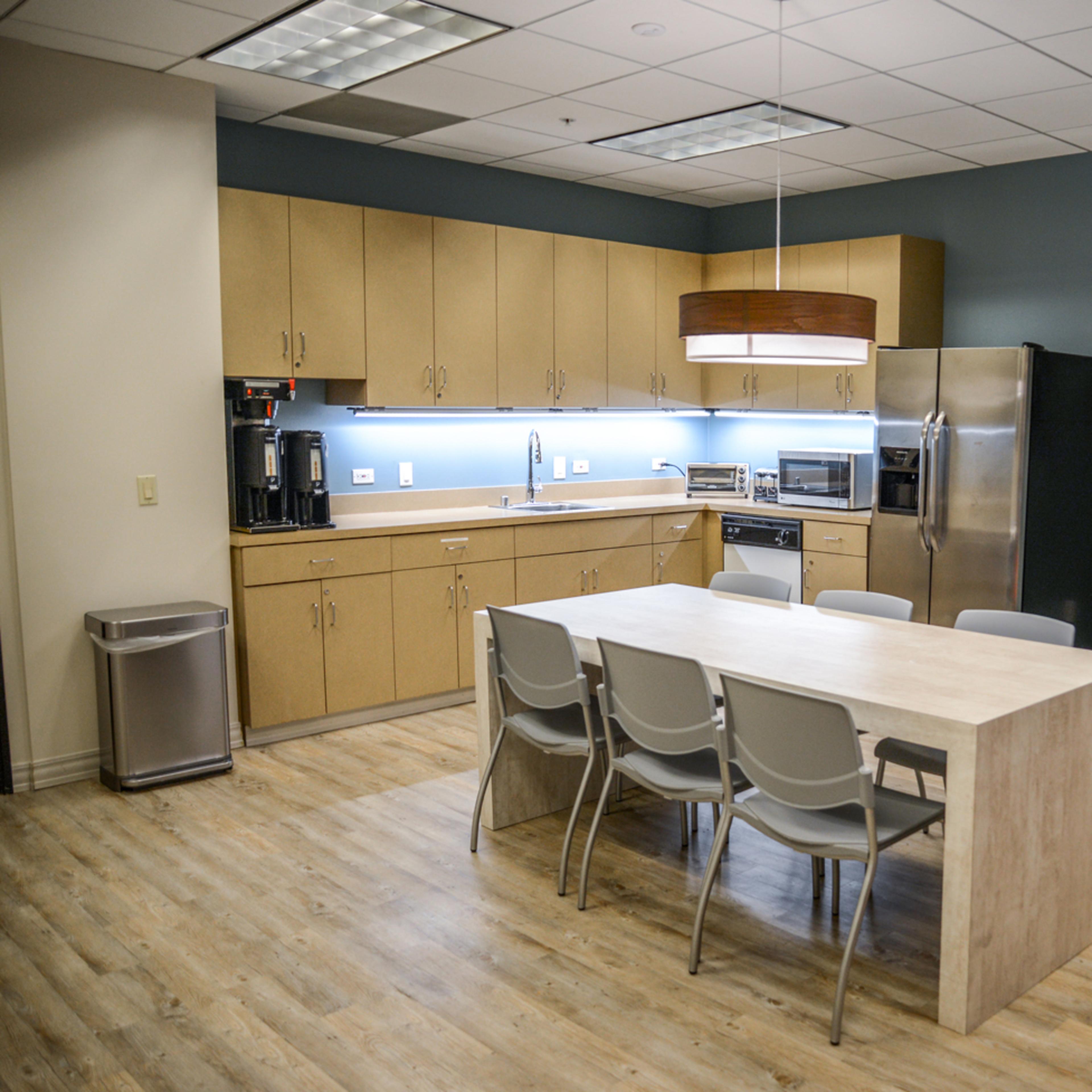 A modern kitchen space with beige cabinets, a stainless steel refrigerator, a dining table surrounded by gray chairs, and a wall-mounted television.