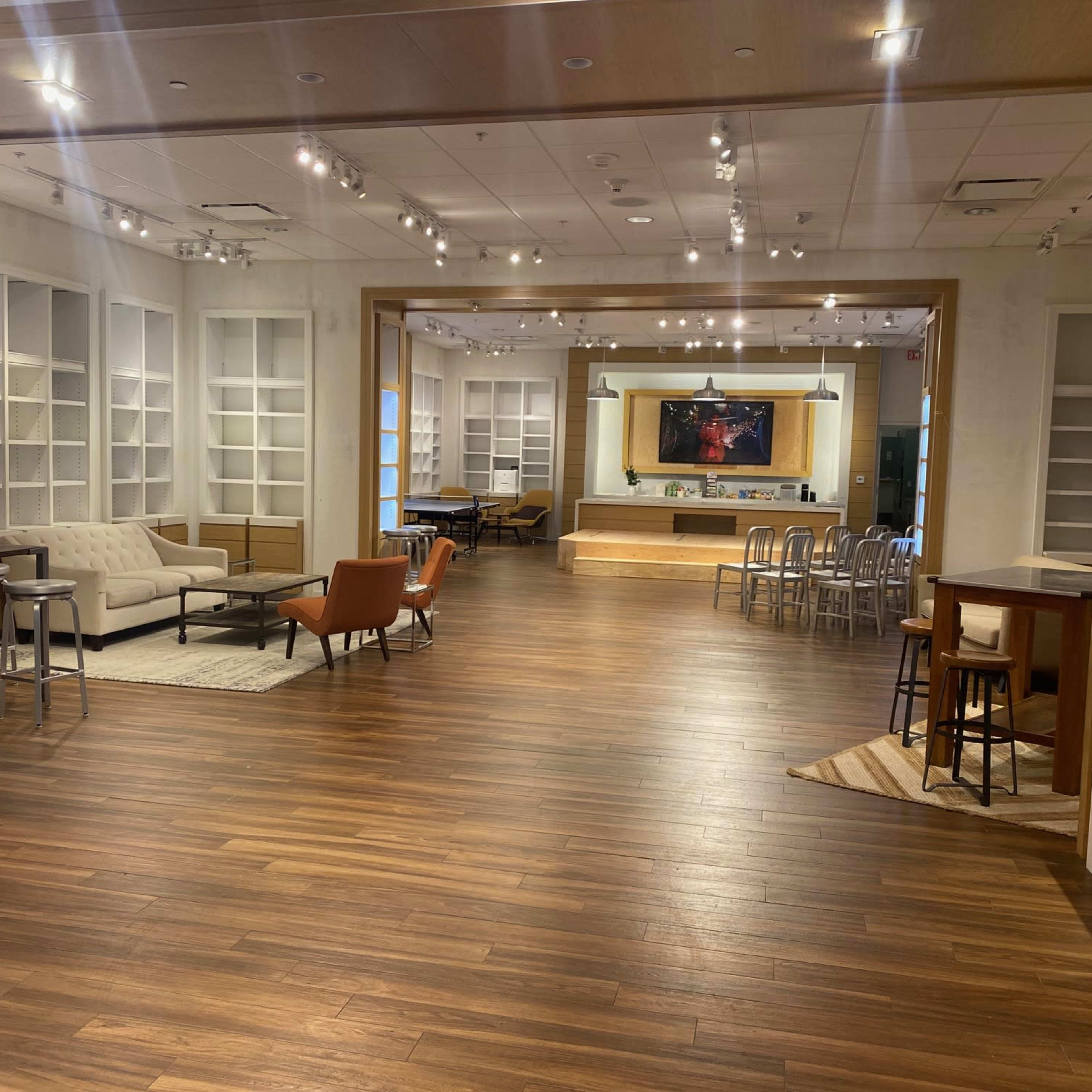 A spacious room with wooden flooring, featuring a seating area with a sofa and armchairs, a kitchen space in the background, and built-in shelves along the walls.