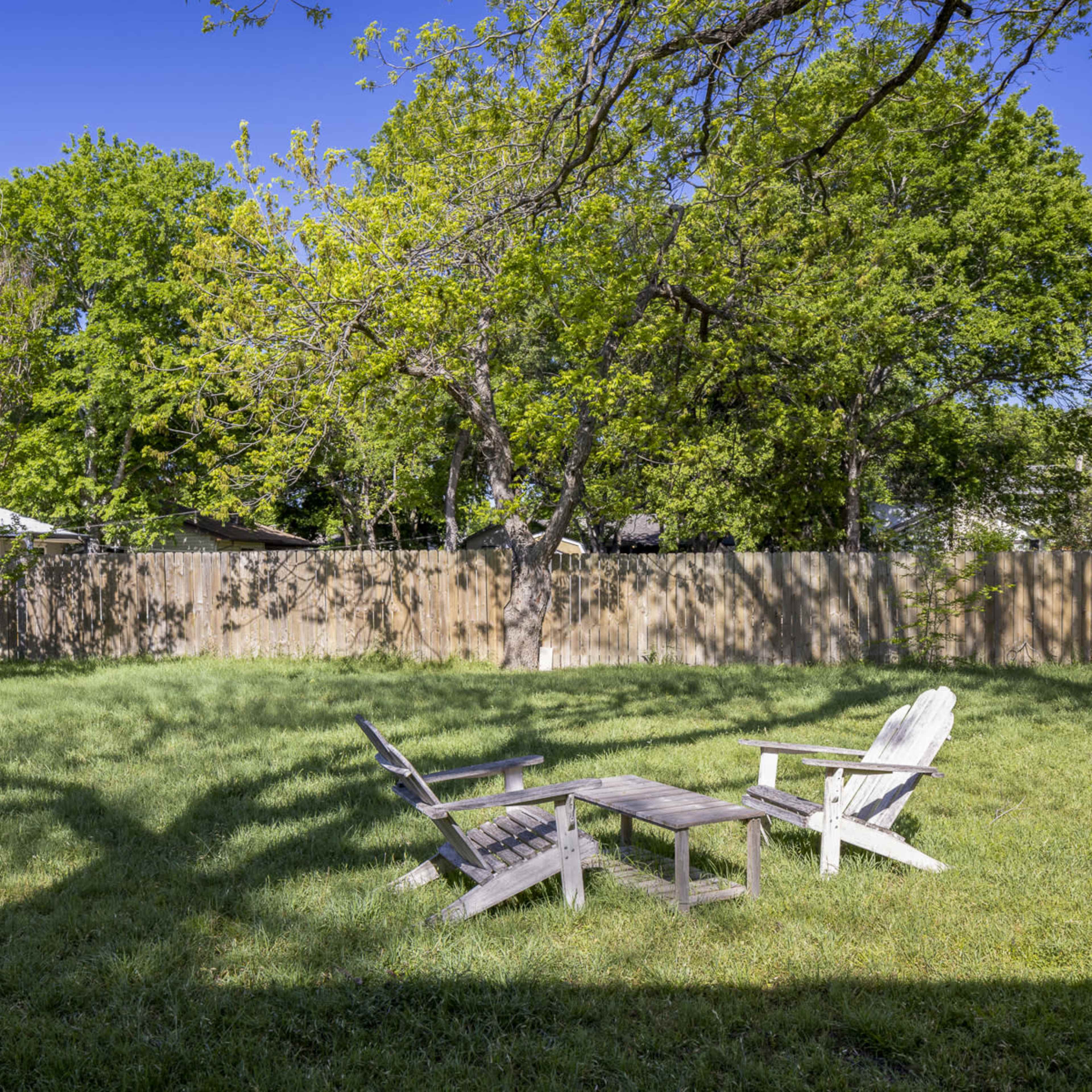 A wooden table and four chairs are arranged in a grassy backyard surrounded by a wooden fence and trees.