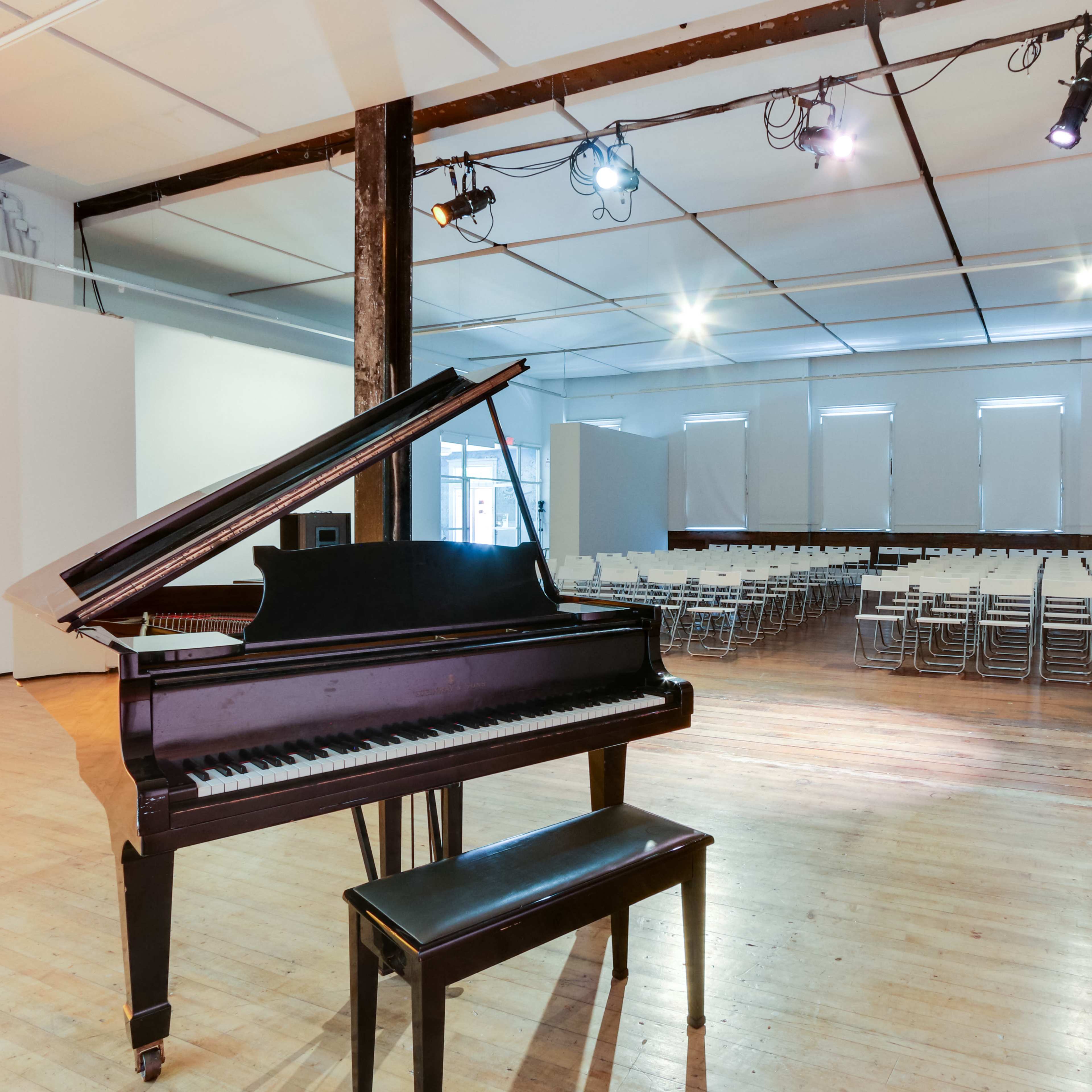 A grand piano and bench are positioned in the foreground of a spacious room set up with rows of chairs for an event.