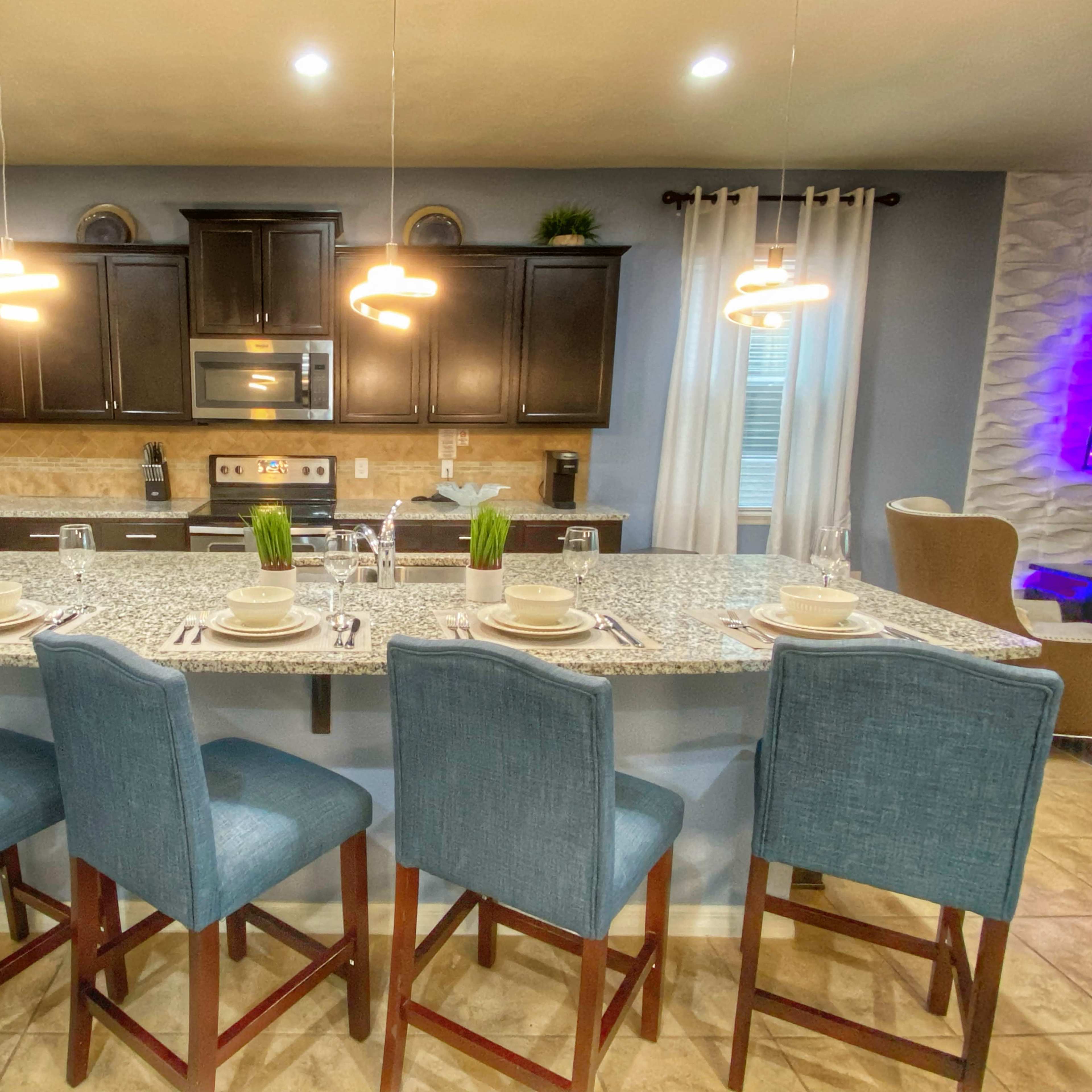 A modern kitchen features a granite countertop island with four blue bar stools, stainless steel appliances, and warm cabinetry.