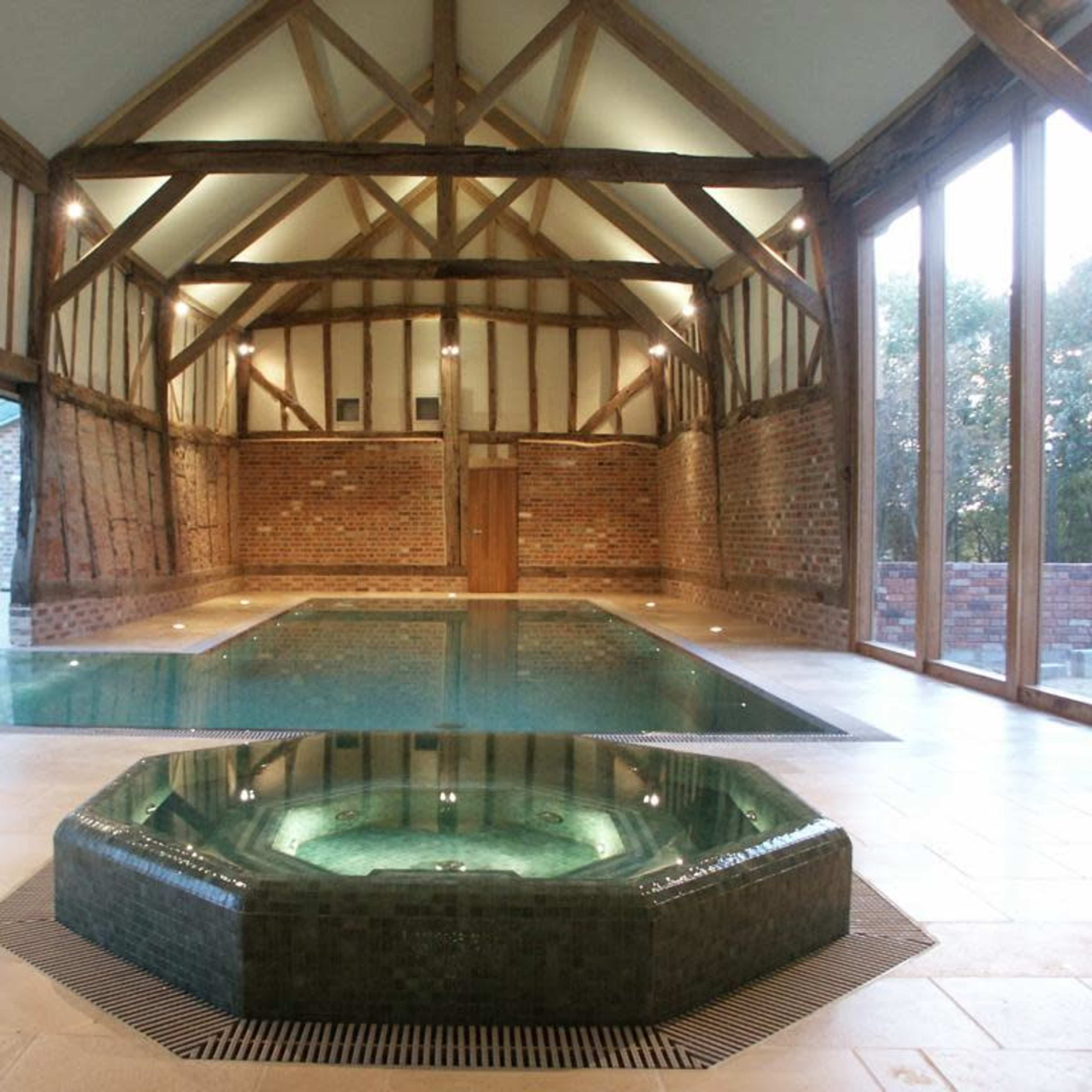 An indoor swimming pool with a sunken hot tub, set in a spacious room featuring exposed wooden beams and large glass windows.