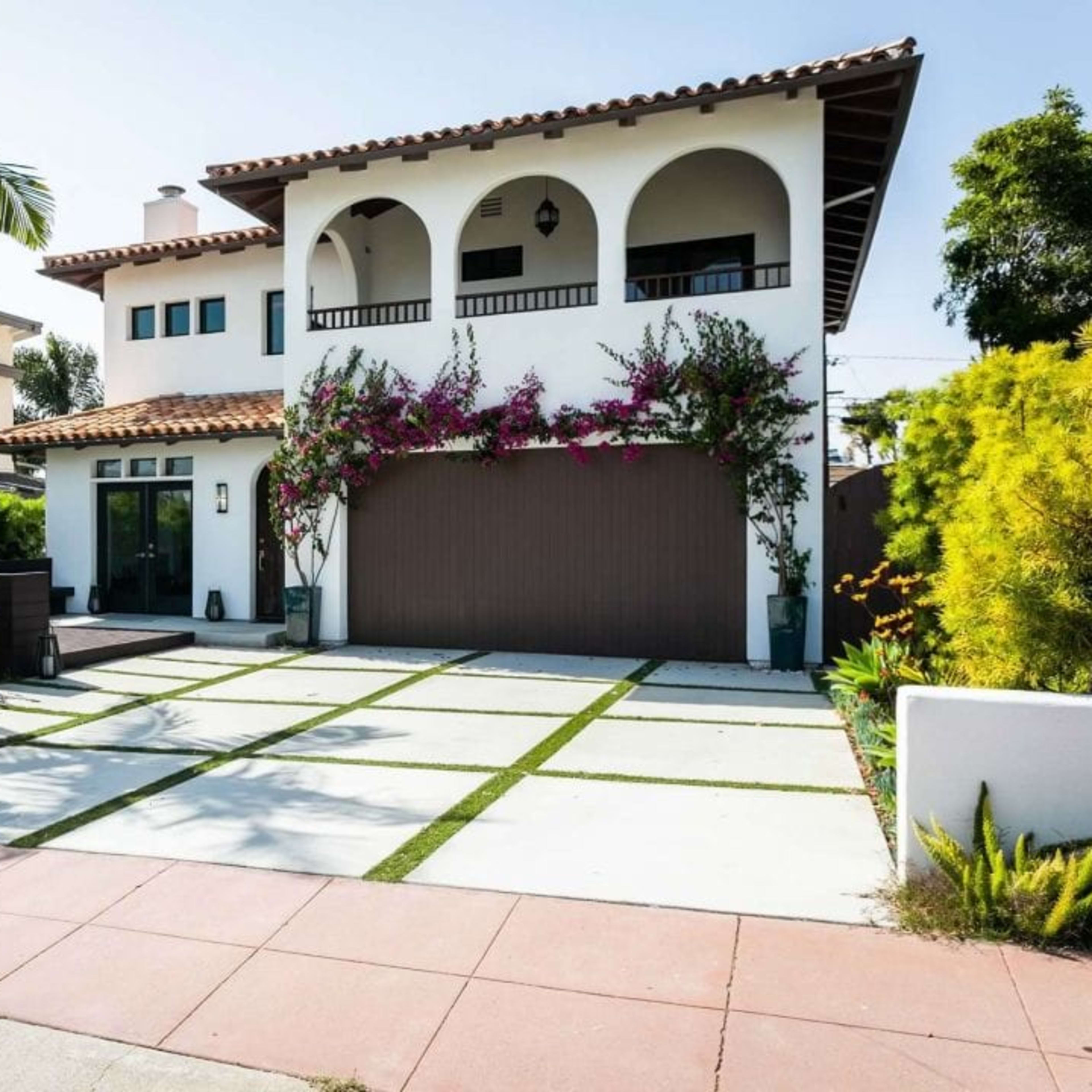 A two-story house with a tiled roof, a large garage door, and a landscaped front yard featuring palm trees and vibrant flowers.