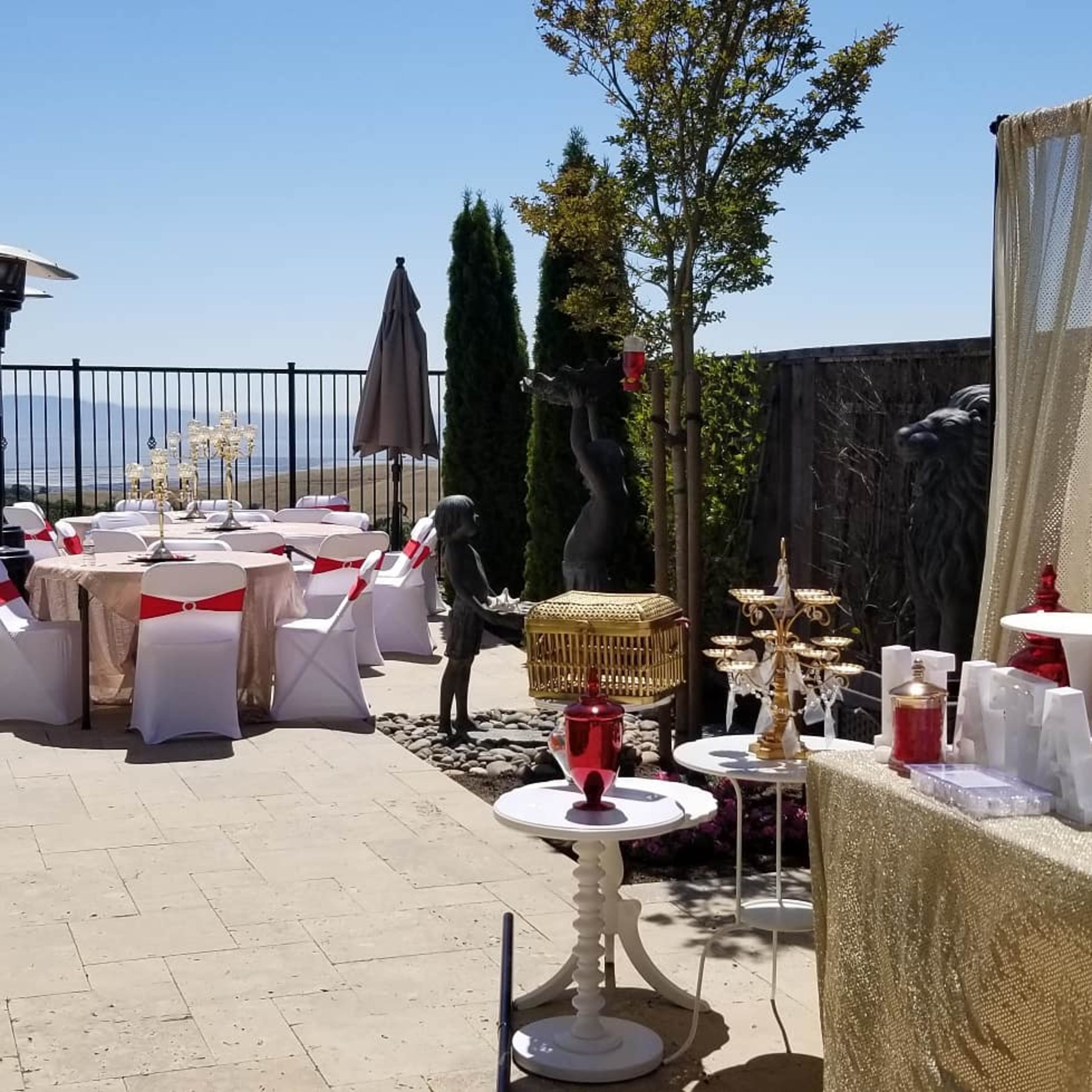 The scene features a terrace setup for an event, with decorated tables, a statue, and lush greenery overlooking a panoramic view.