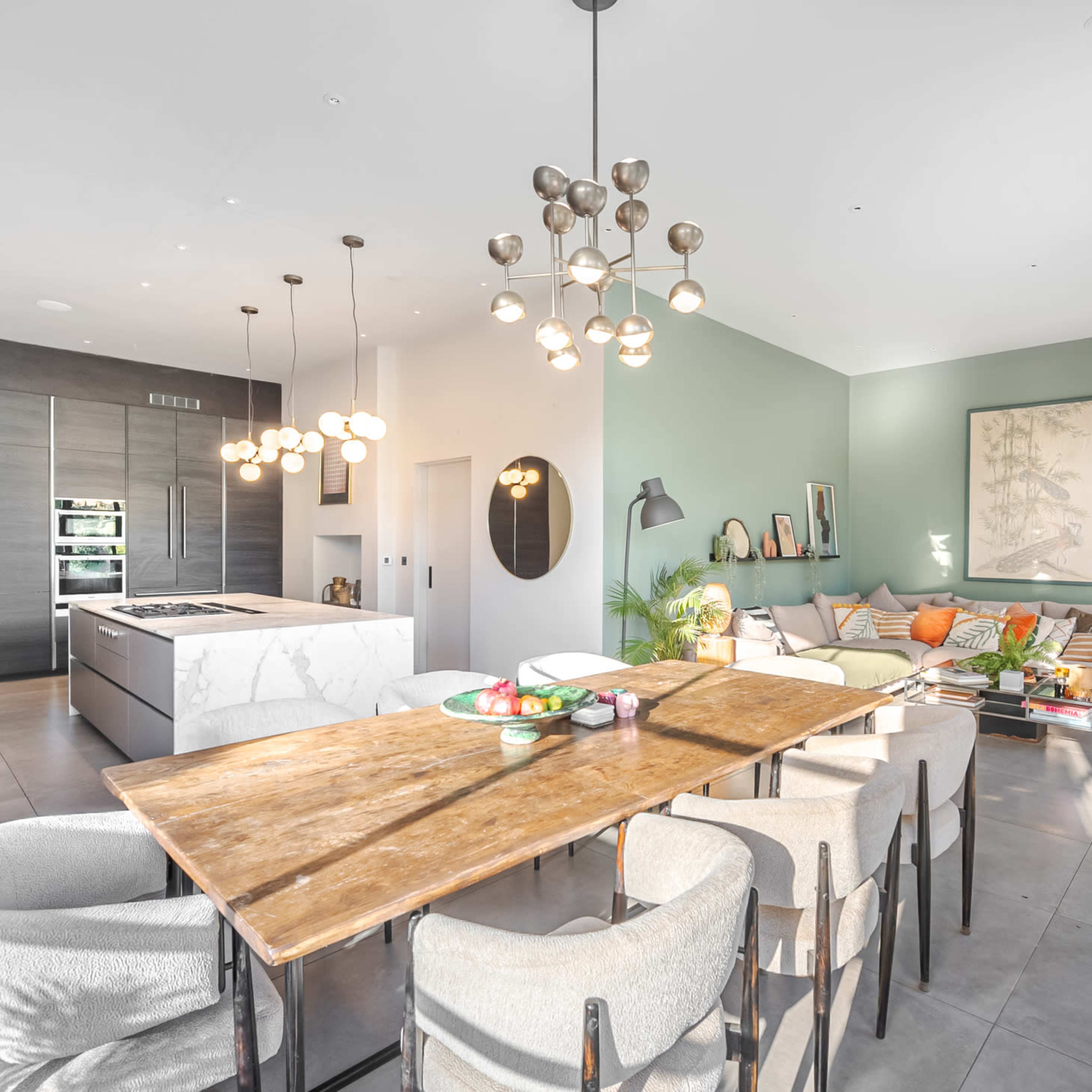 A modern kitchen and dining area featuring a wooden table surrounded by upholstered chairs, a marble kitchen island, and a seating area with a sofa and decorative plants.