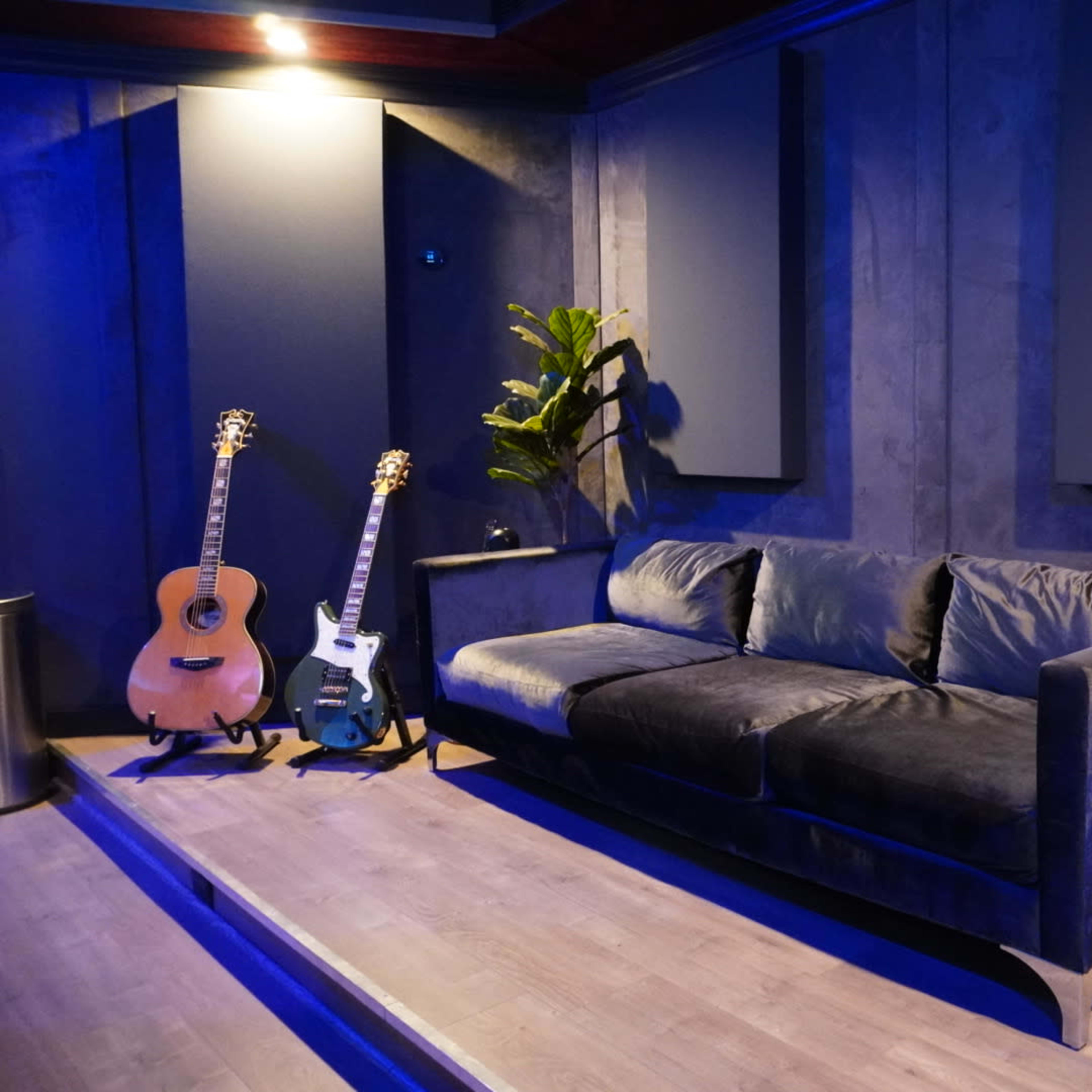 A modern music studio featuring two guitars, a dark sofa, a vintage fridge, and a potted plant against a backdrop of blue lighting.
