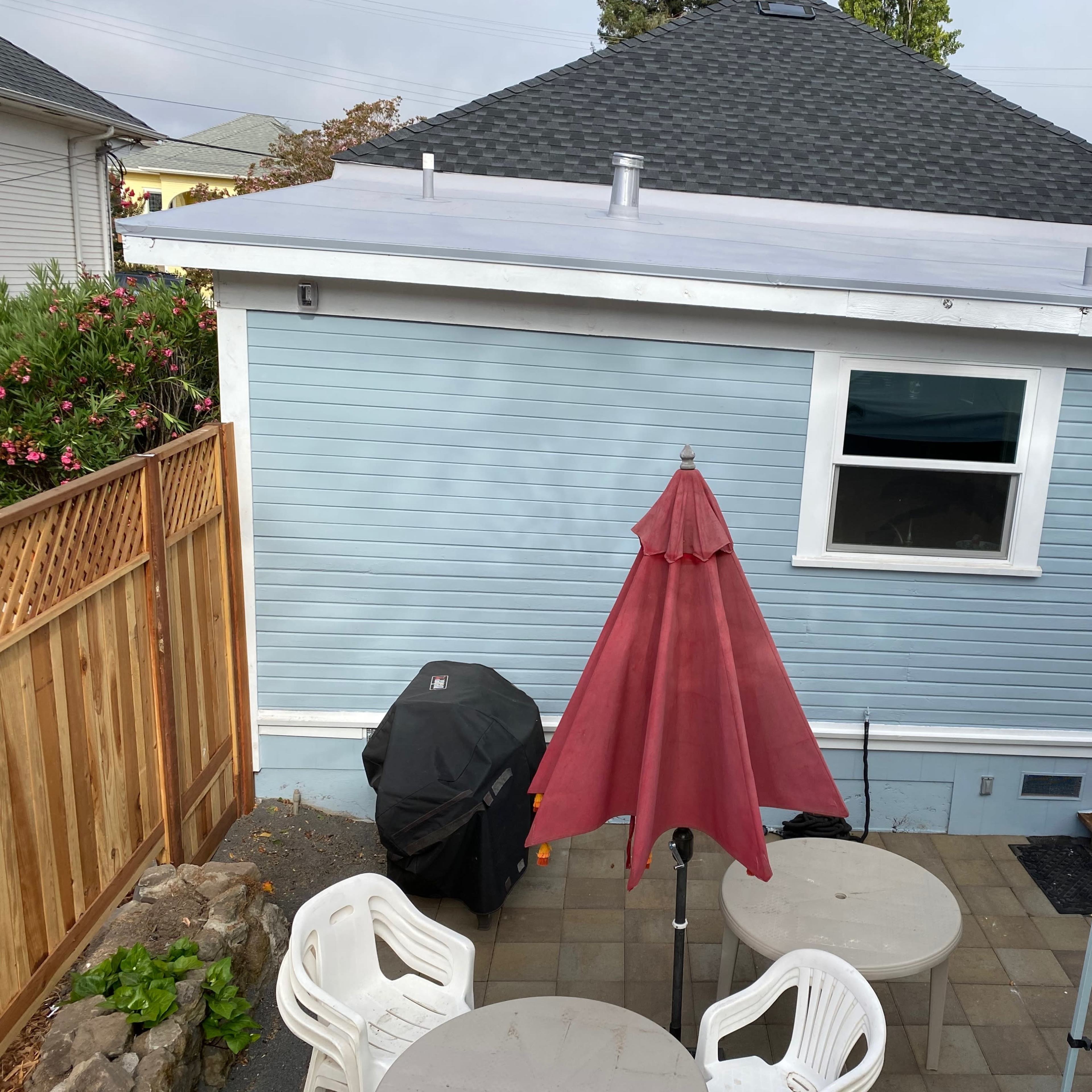A backyard scene features a light blue house with a red umbrella over two white tables, a grill, and a stone patio.