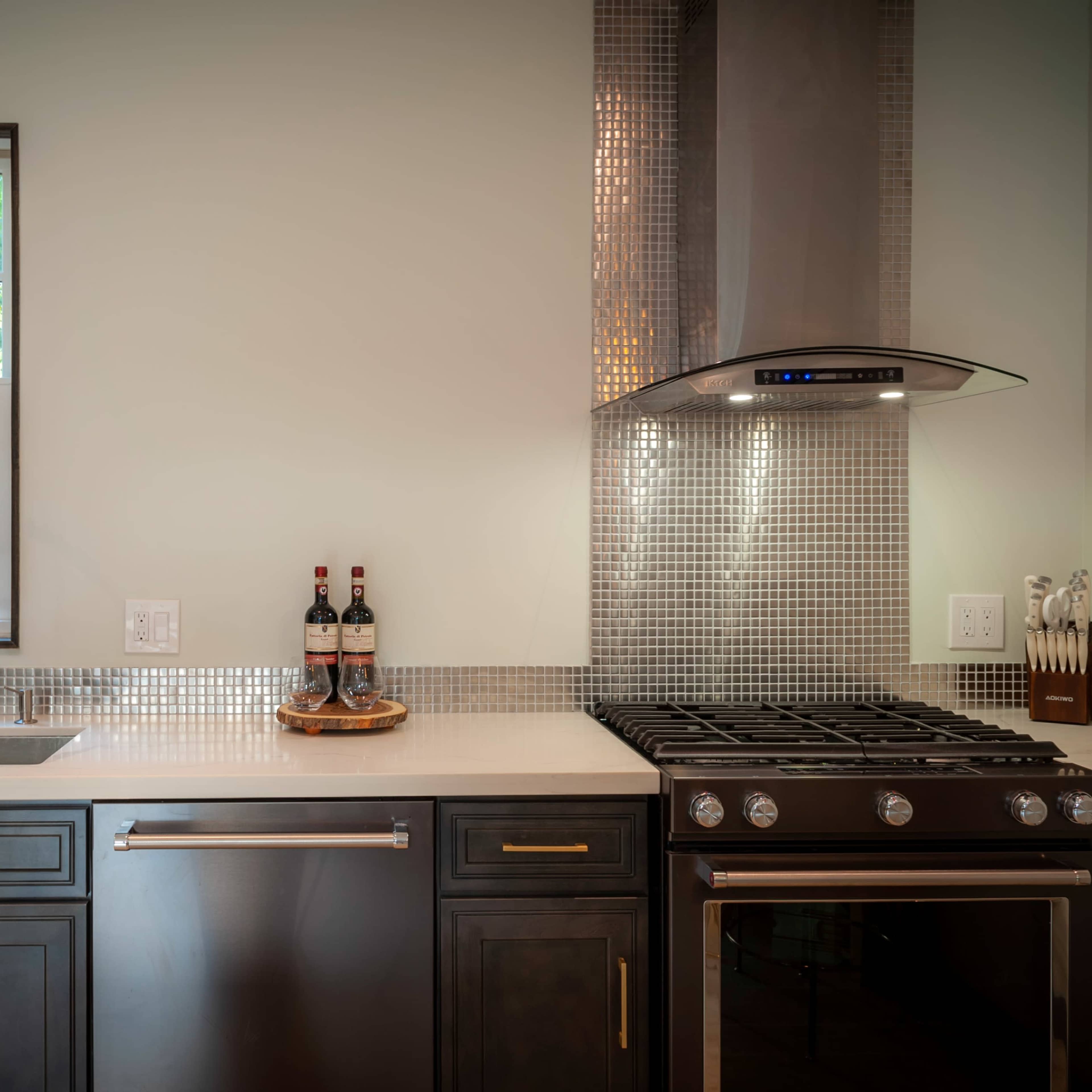 A modern kitchen features a stainless steel sink, a gas stove, and a mosaic tile backsplash with a window above the sink.