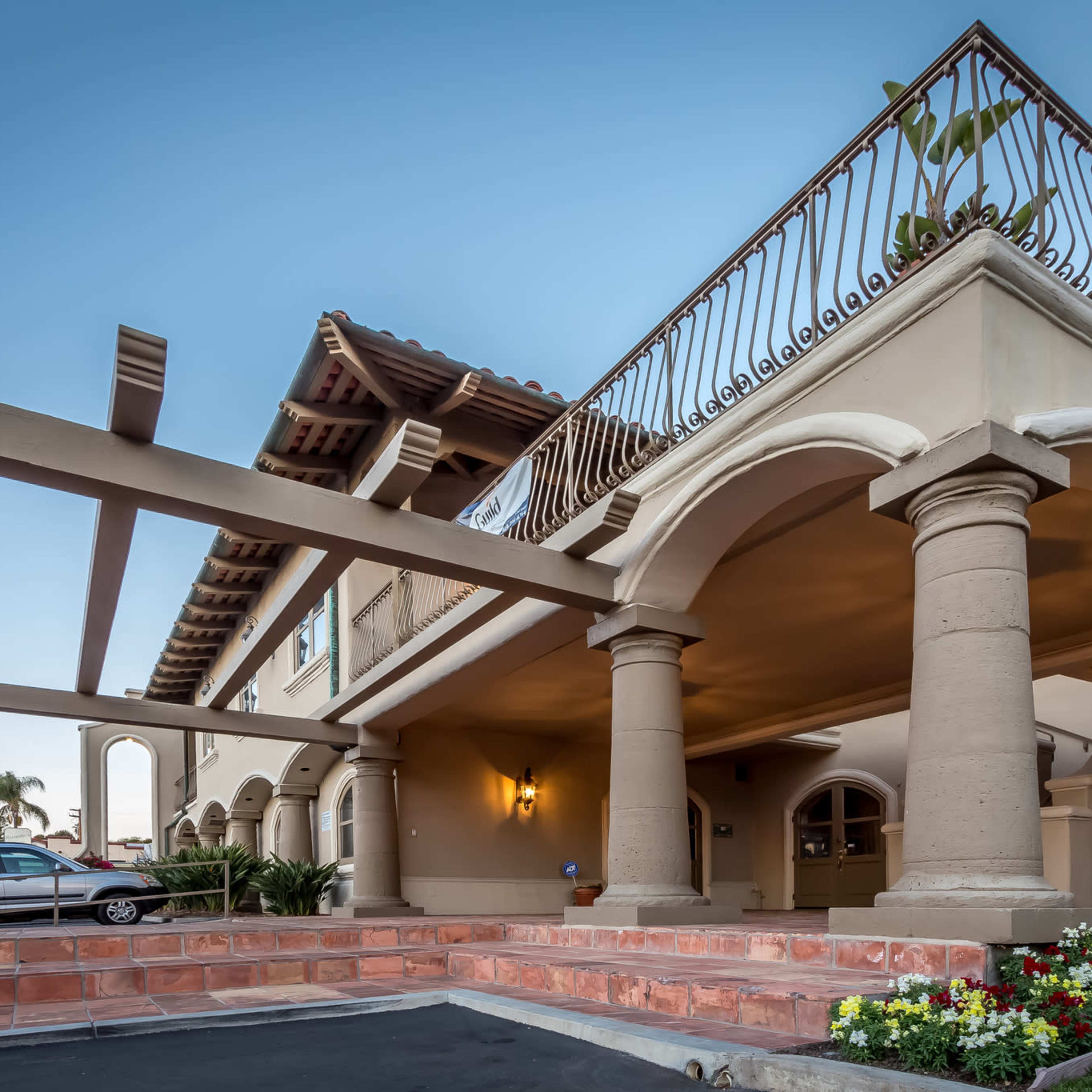The entrance of a building featuring arched columns and a balcony above, with a vehicle parked nearby and flower beds in the foreground.