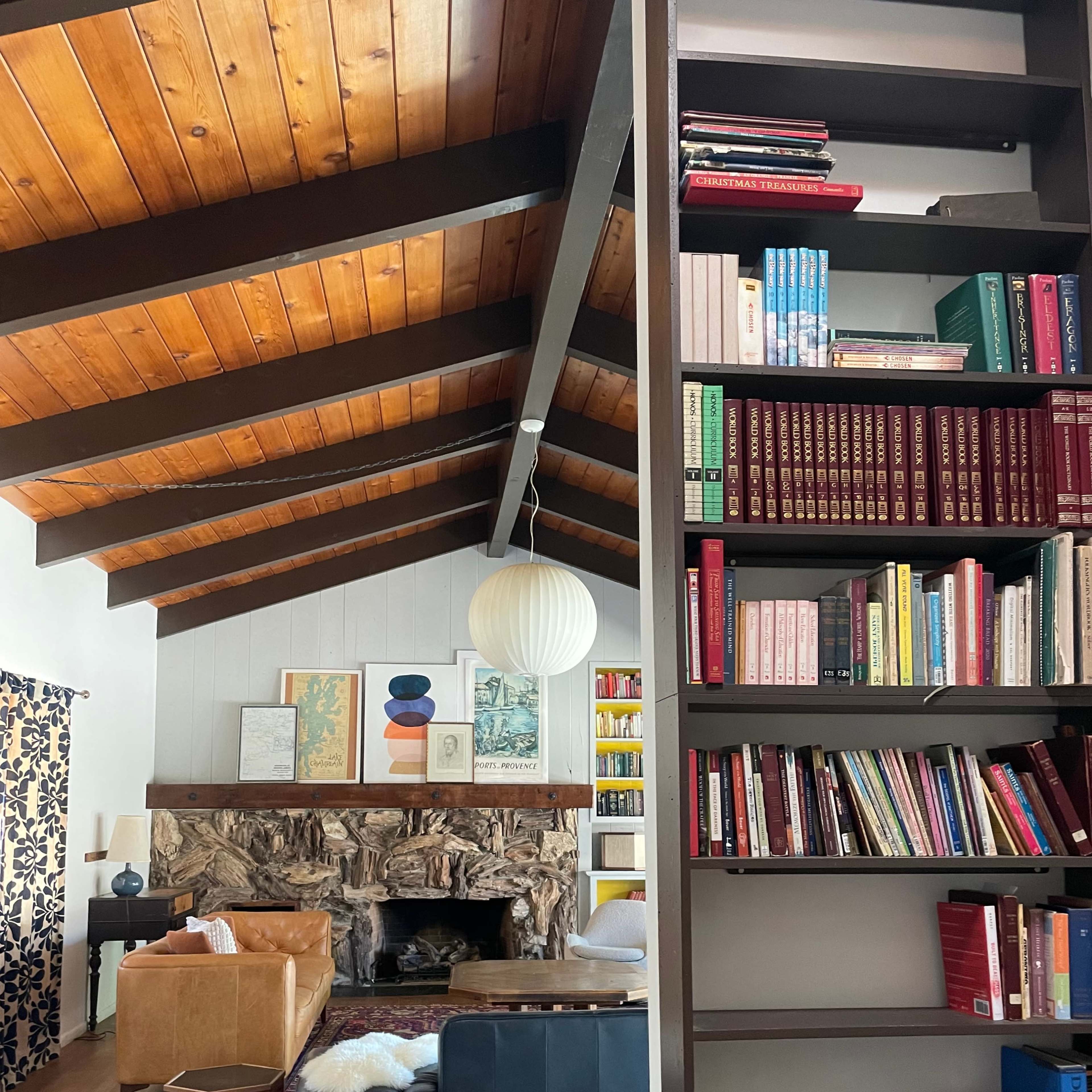 A living room with a high, wooden-beamed ceiling, a stone fireplace, and bookshelves filled with various books.